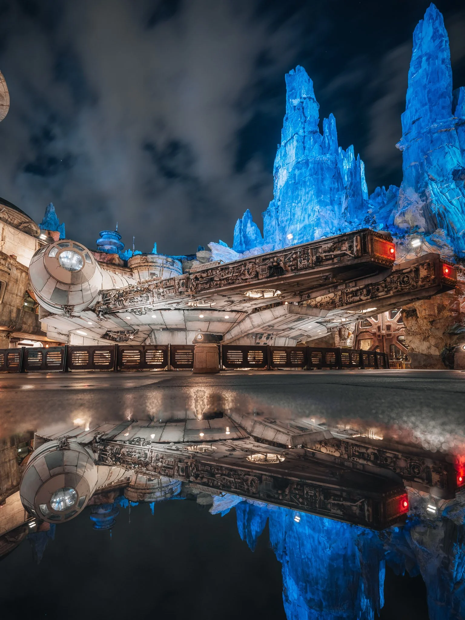 View of a futuristic spaceship docked in front of a tall, glowing blue ice mountain at night, with clouds and a starry sky overhead, reflected on a calm water surface.