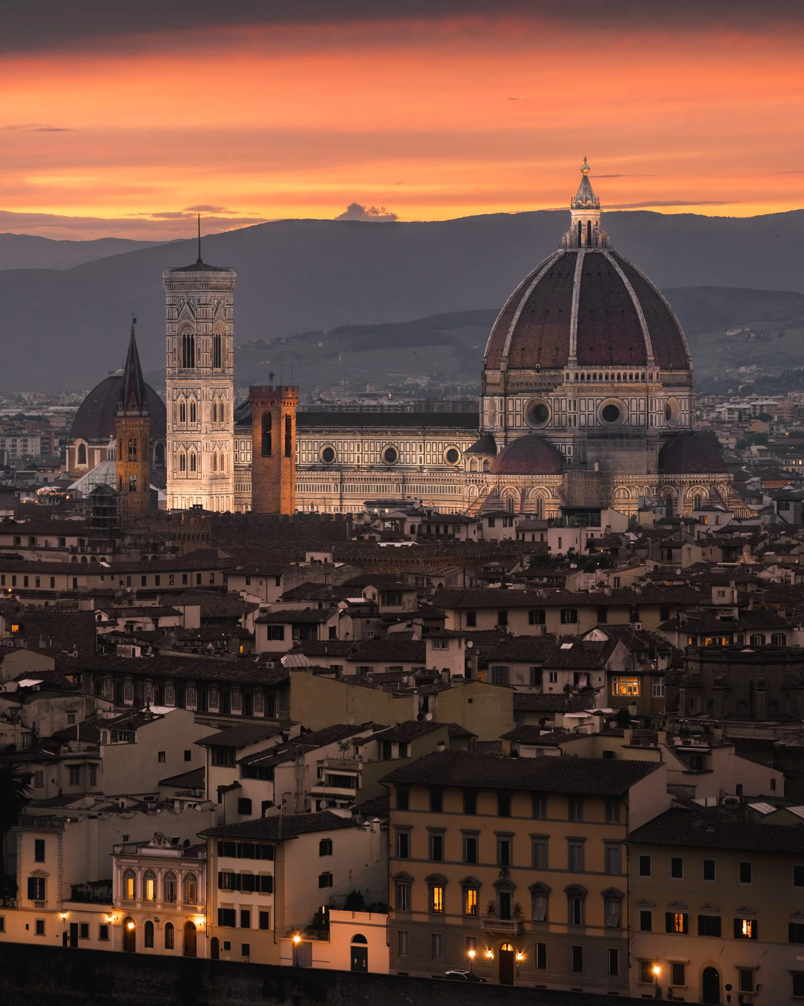 Scenic view of Florence, Italy during sunset, featuring the Florence Cathedral's dome and bell tower surrounded by residential buildings with lit windows, with hills in the background.