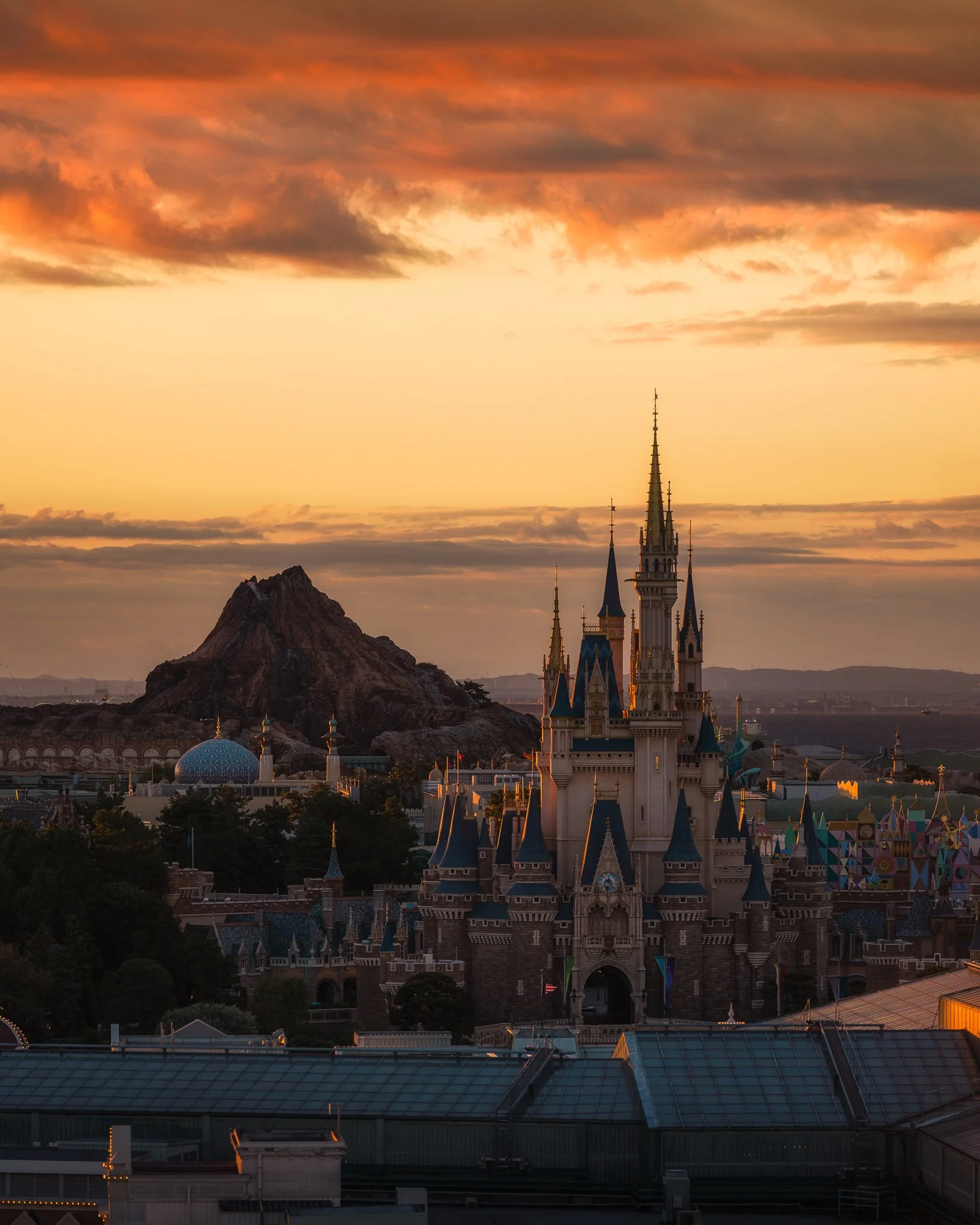 A castle with multiple spires, a blue domed building, trees, and a mountain in the background during sunset with an orange sky and clouds.