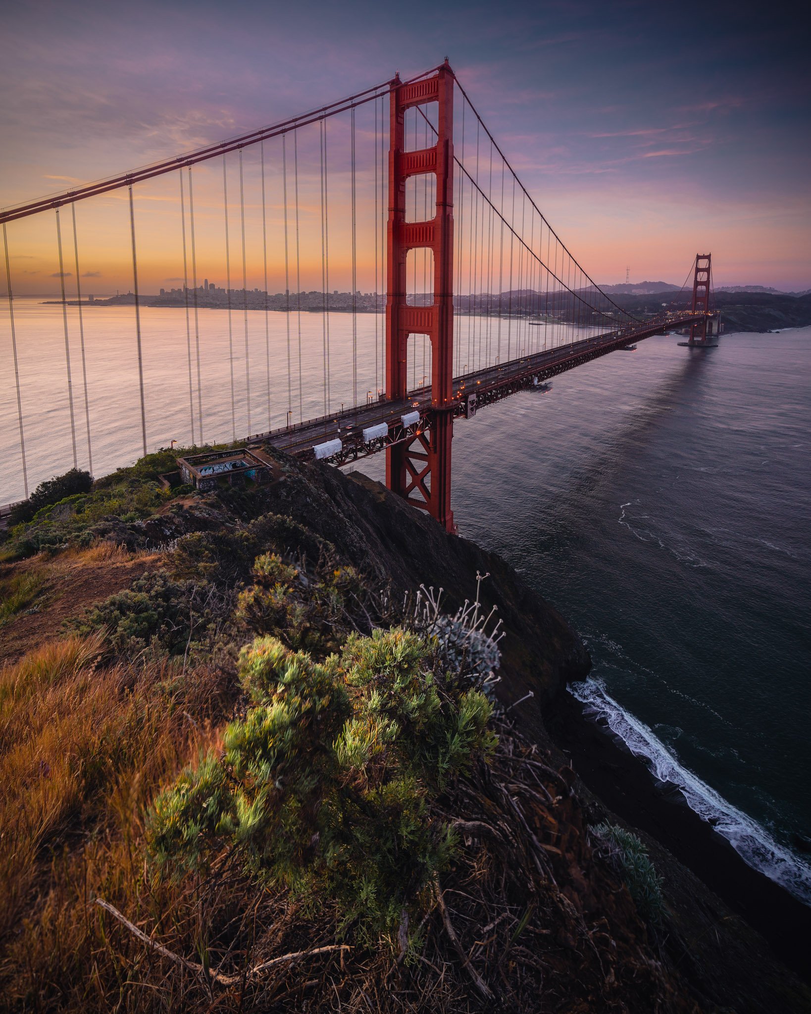 Golden Gate Bridge during sunset over the water, with the city skyline in the distance and a rocky, grassy hillside in the foreground.