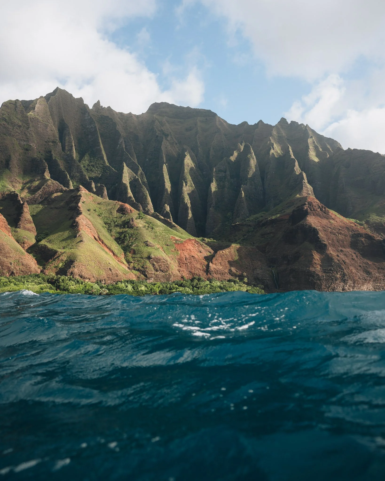Ocean water with waves, green and brown rocky mountainous shoreline with lush vegetation, partly cloudy sky.