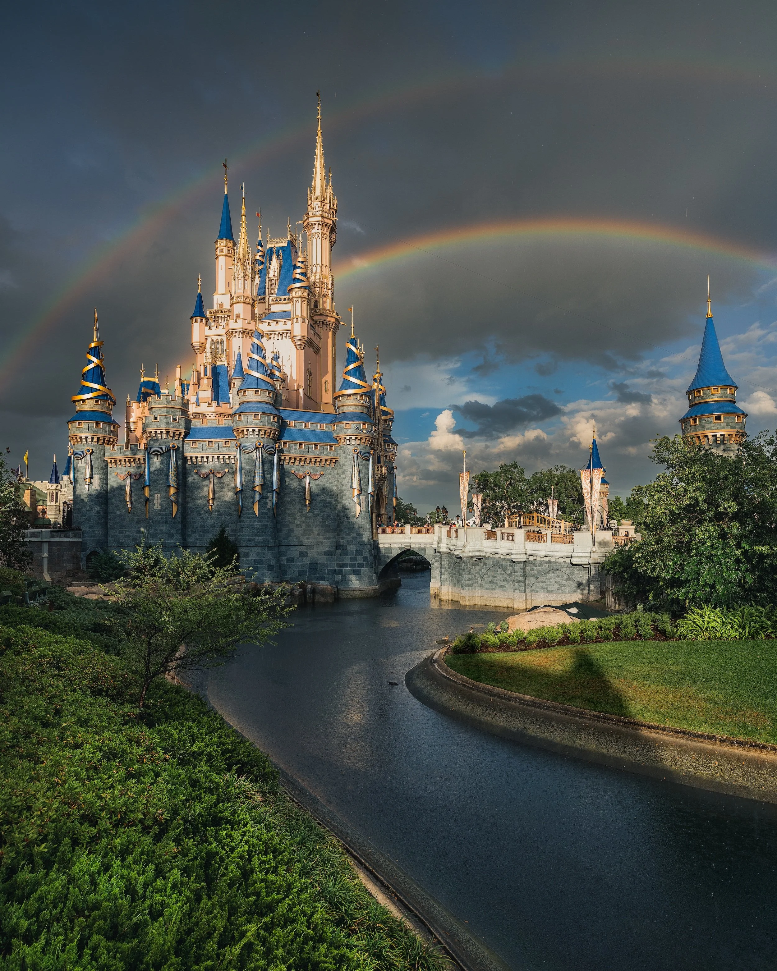 Disney castle with blue rooftops and golden spires, surrounded by greenery, with a rainbow in a partly cloudy sky.