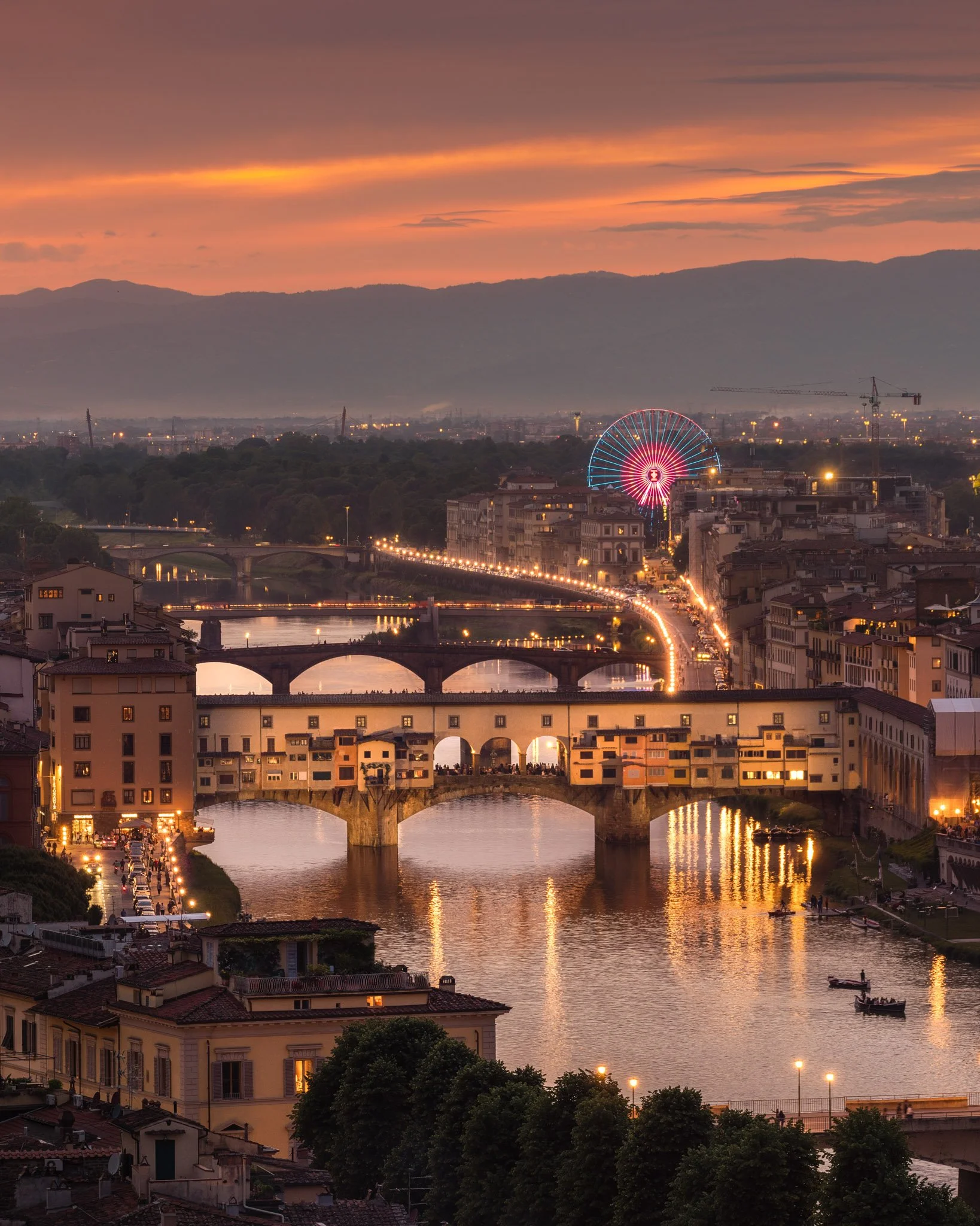 Sunset over a city with a river and a bridge, featuring a large illuminated Ferris wheel, residential buildings, trees, and a mountainous background.