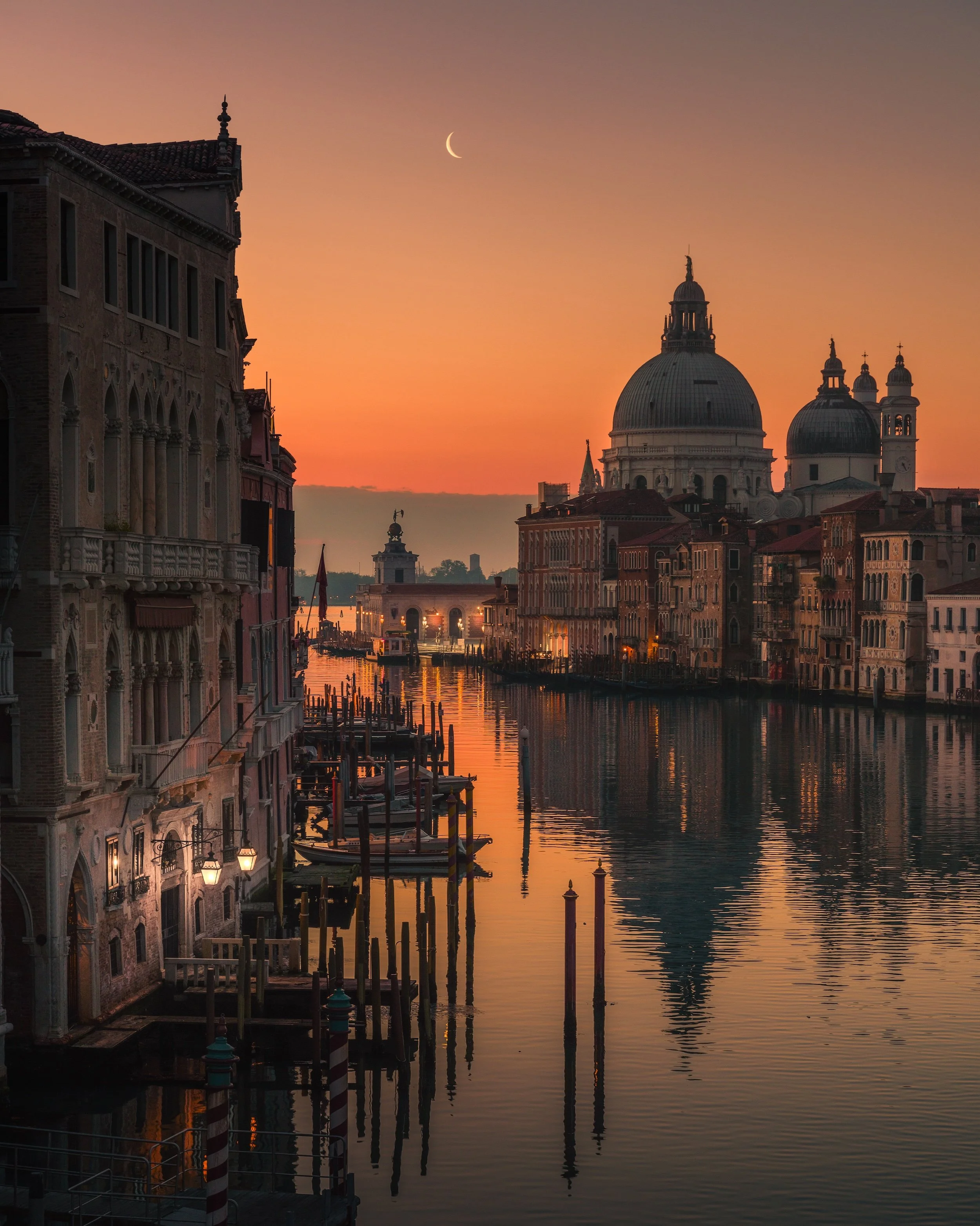Sunset over a canal in Venice, Italy, with historic buildings, a crescent moon in the sky, and boats docked along the water.