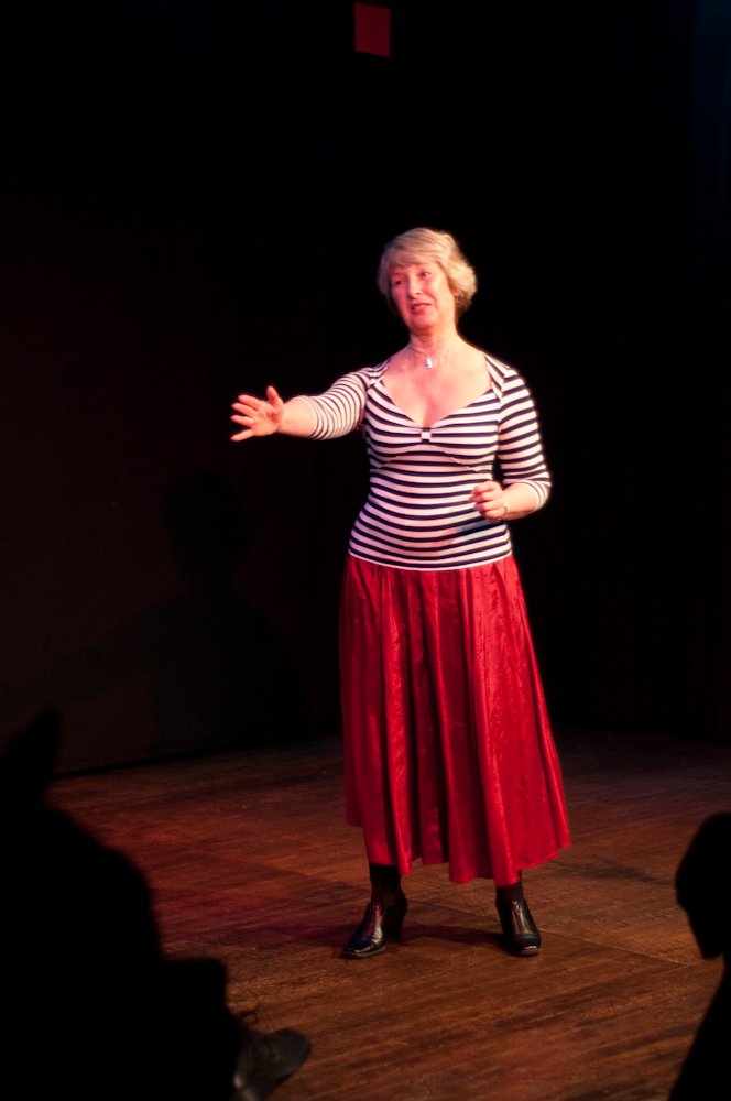 A woman performing stand-up comedy on stage, wearing a striped shirt and red skirt.