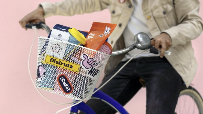 Person on a bicycle with a white basket containing various colorful bags and containers, decorated with stickers against a pink background.
