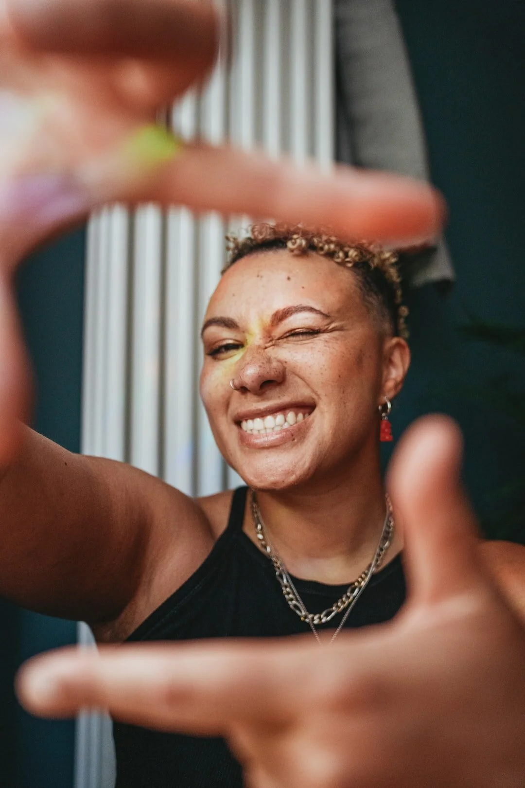 Person smiling and framing their face with their hands, wearing a black top and jewelry, squinting one eye.