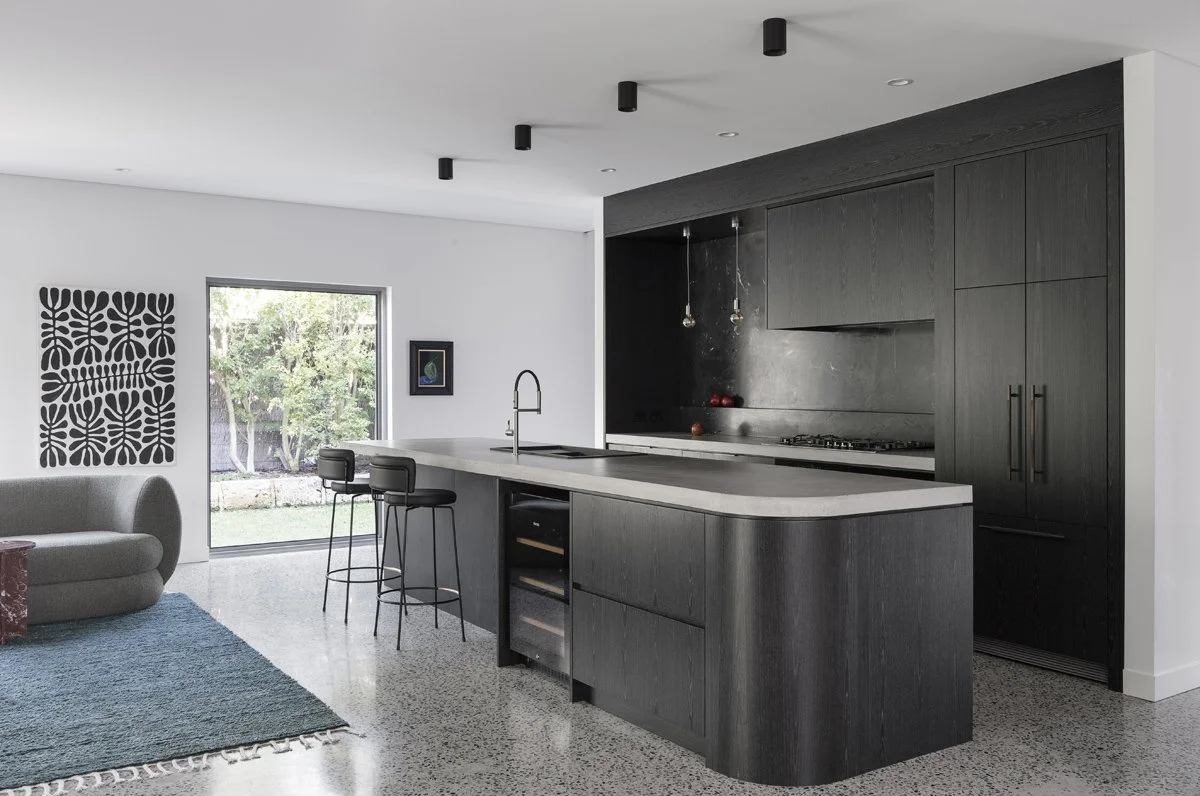 Modern kitchen with black cabinets, dark wood island with white countertop, black bar stools, and a sliding glass door leading to a yard.