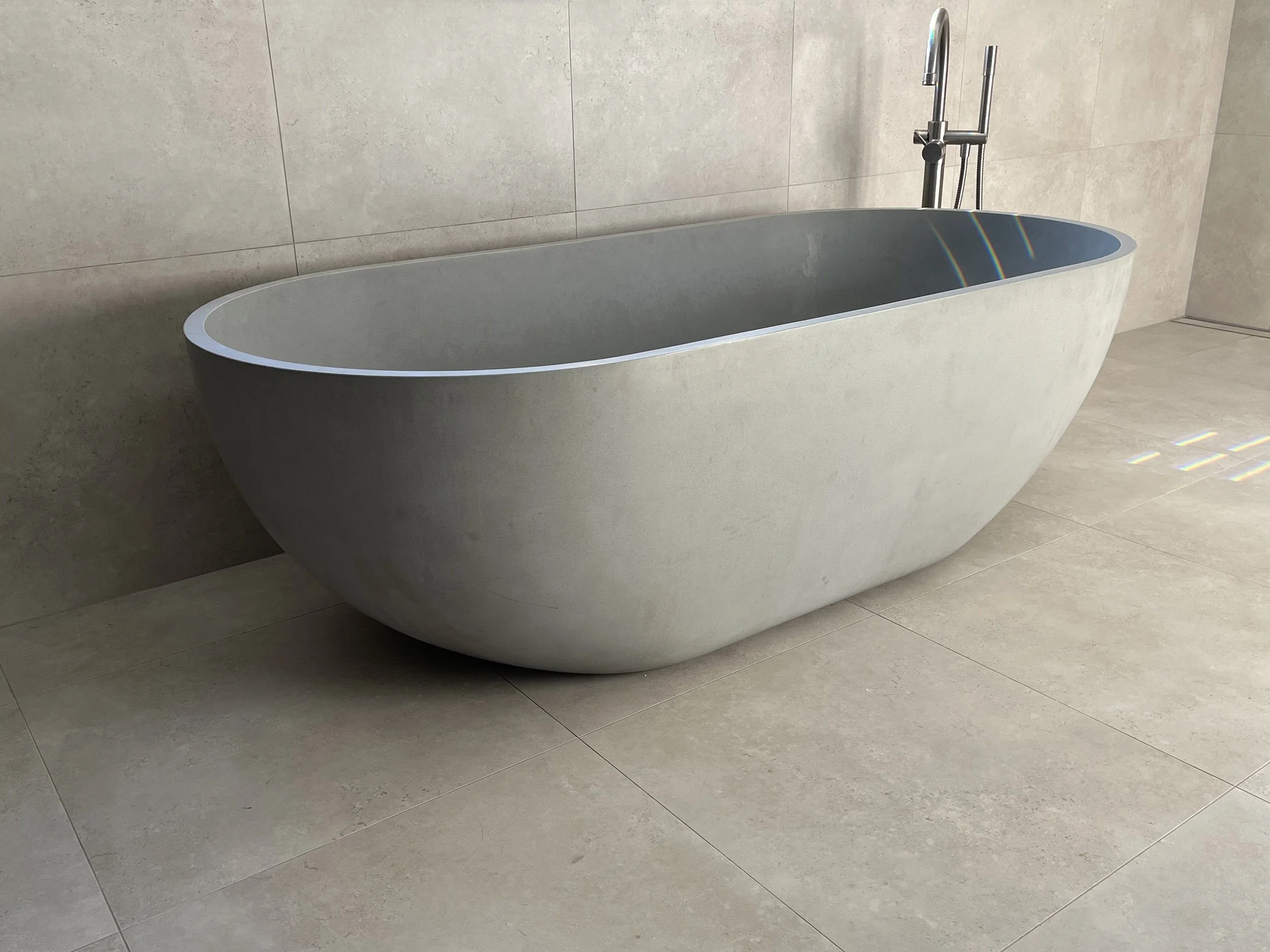 Modern freestanding oval bathtub with a chrome floor-mounted faucet against a beige tiled wall and floor.