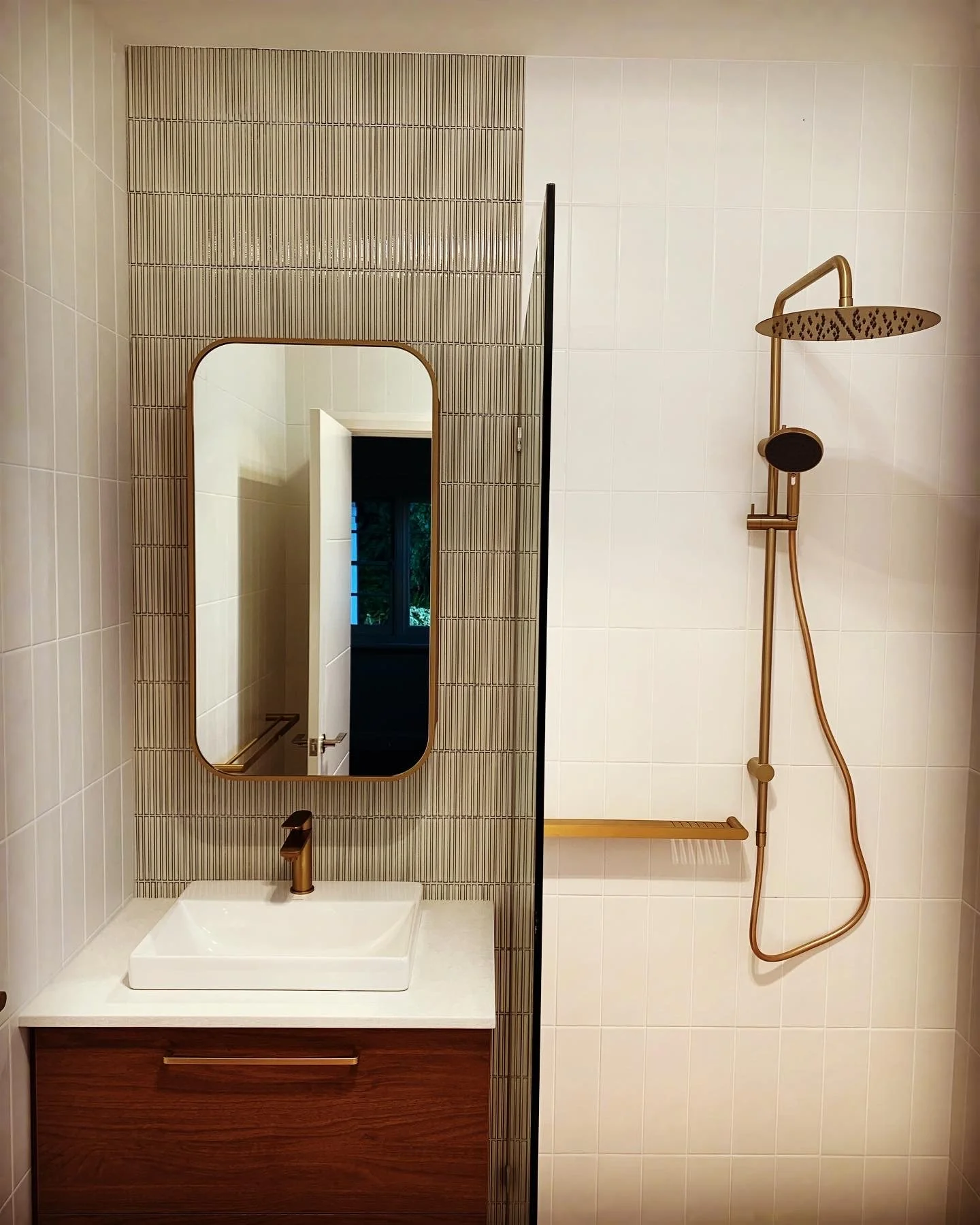 Modern bathroom with a white sink, rectangular mirror, and a wooden vanity. To the right, there is a gold-colored showerhead with a handheld attachment and a wooden shelf.