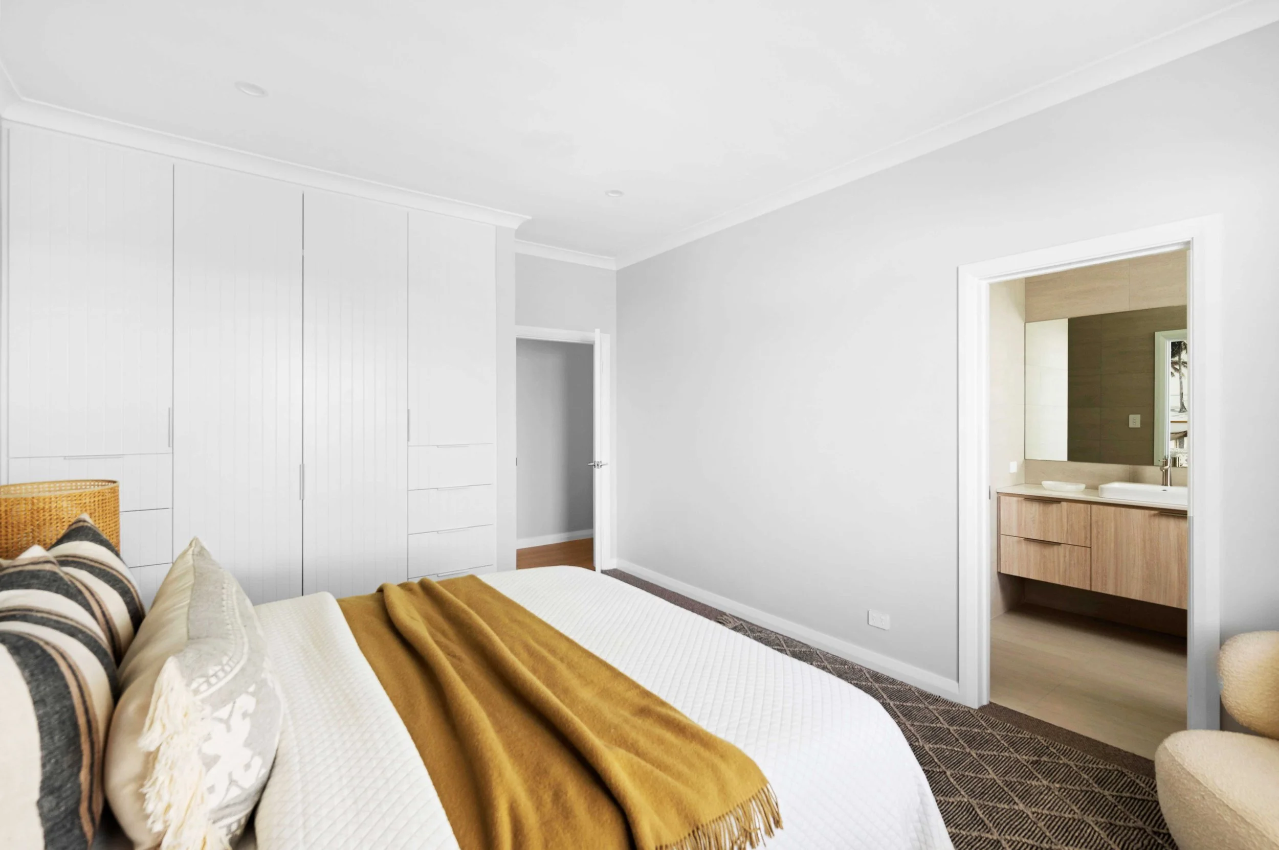 A bedroom with white walls, a bed with striped and cream pillows, a mustard yellow blanket, built-in white closet, and a doorway leading to a bathroom with a wooden vanity and mirror.