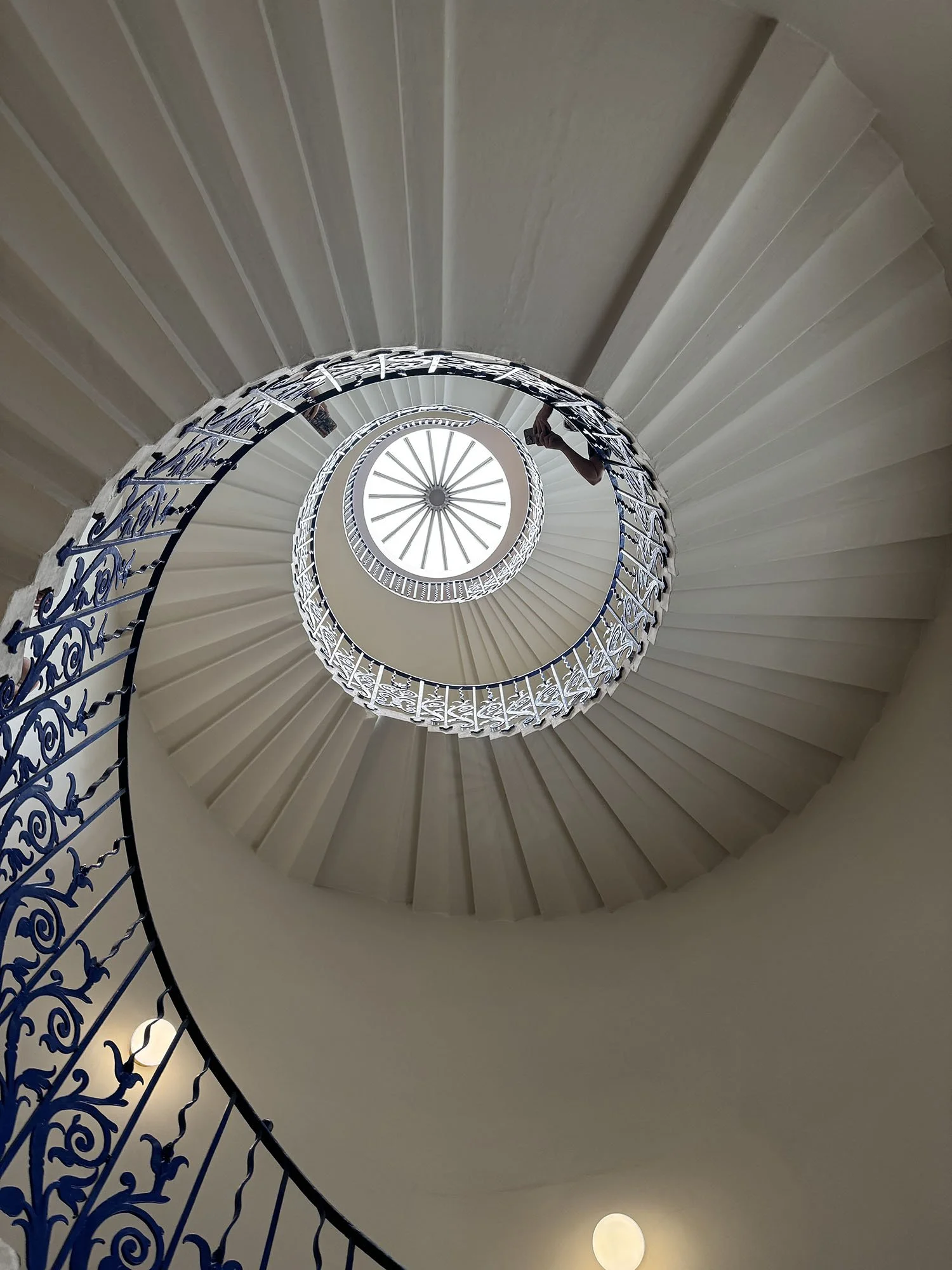 Spiral staircase viewed from the bottom looking up, with white steps, an ornate wrought iron railing, and a circular window at the top letting in sunlight.