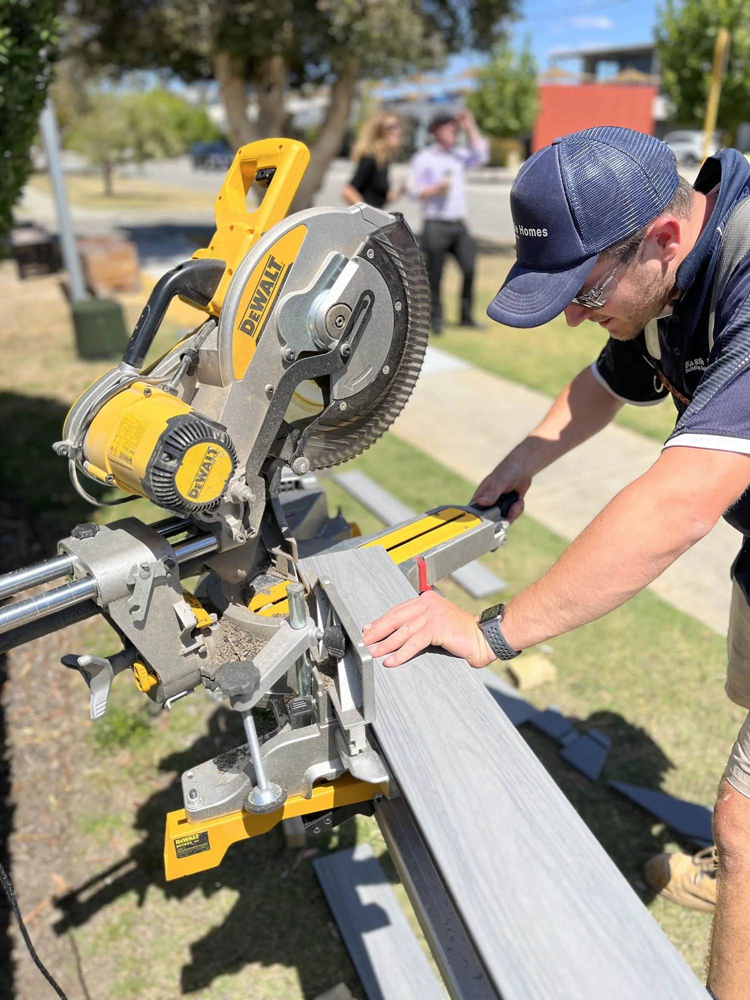 A man operating a DeWalt miter saw outdoors, cutting a wooden board, with two women standing in the background near trees and a street.