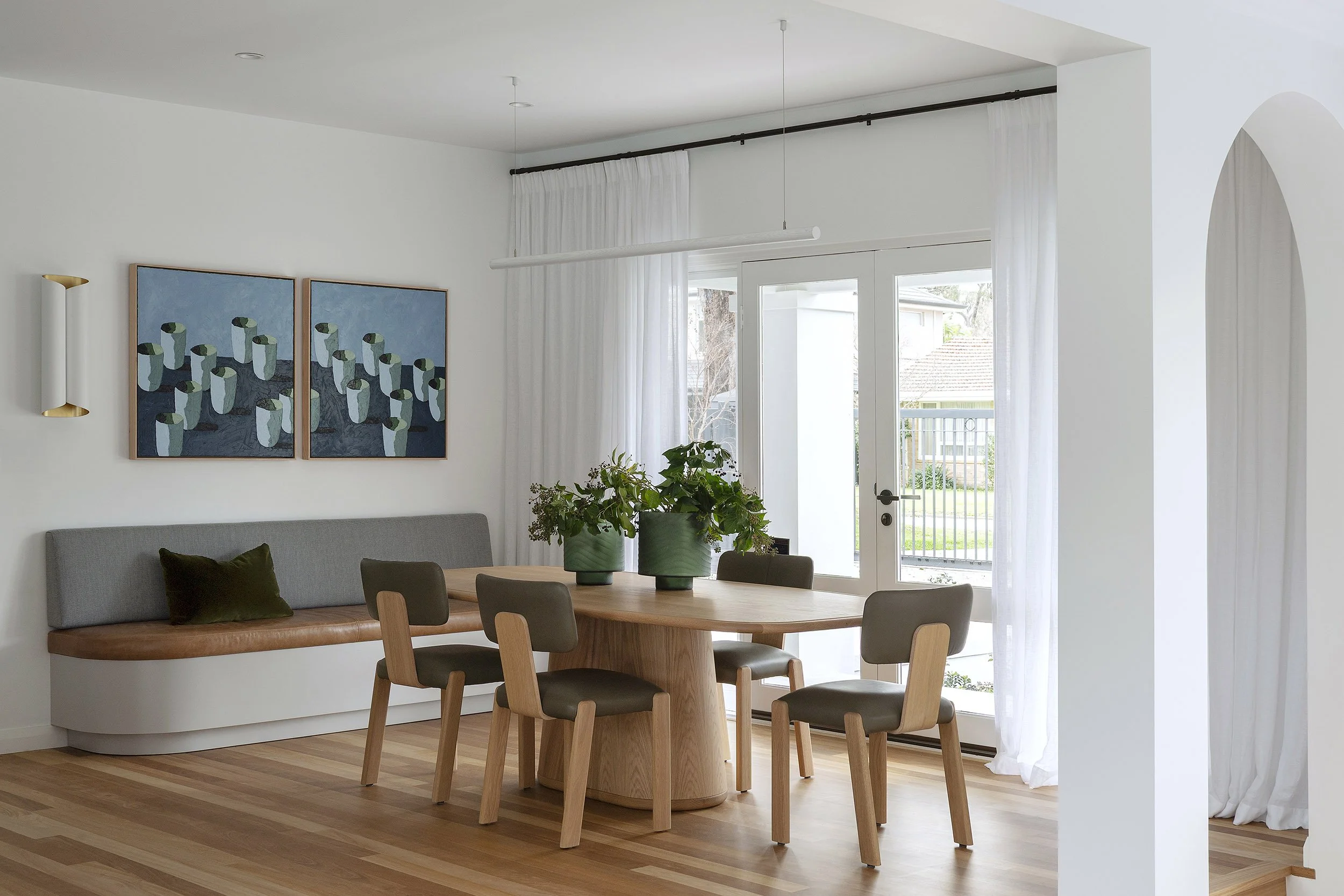 Modern dining area with a wooden table, six chairs, green potted plants, a gray bench with a dark green pillow, artwork on the white wall, and white curtains overlooking a backyard.