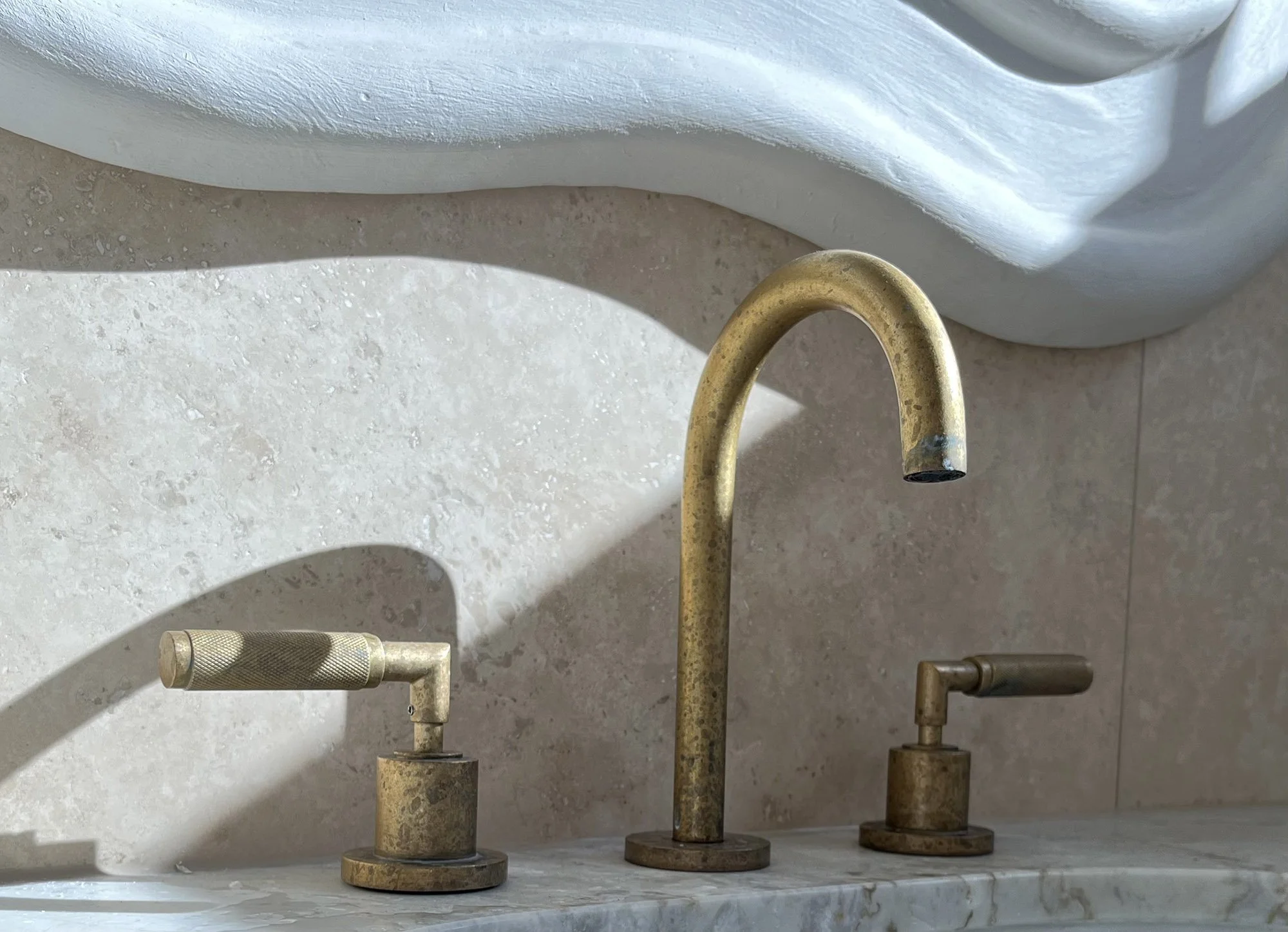 Close-up of a vintage brass faucet with two handles on a marble sink, with a white curved wall in the background.