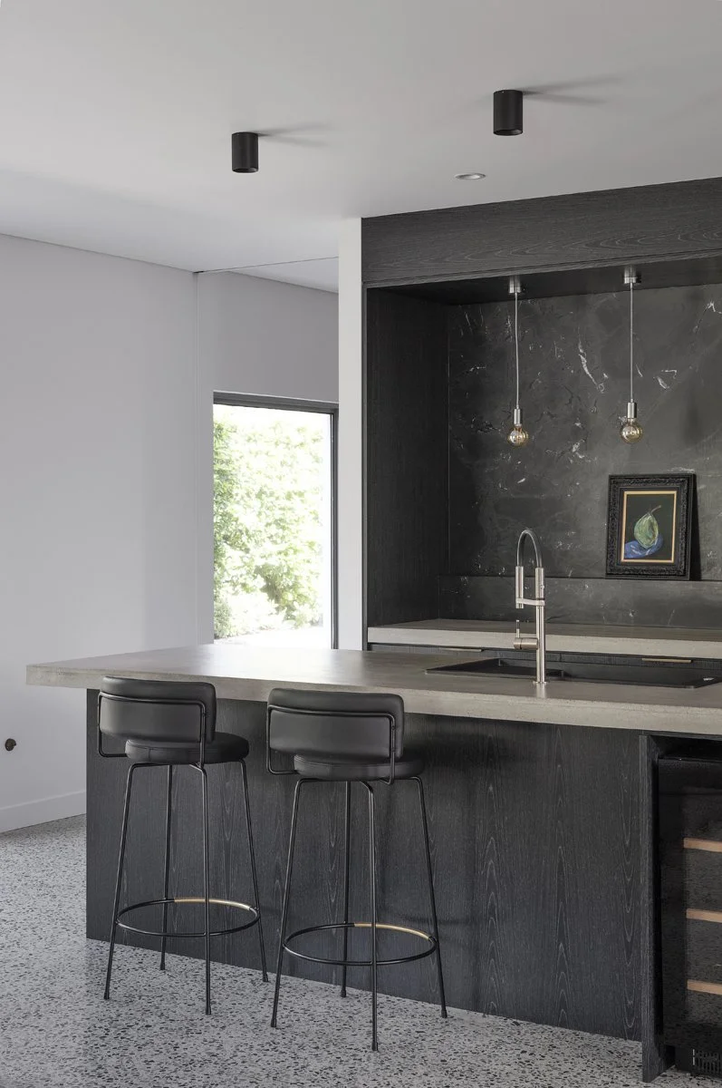 Modern kitchen with black cabinets, marble countertop, two black bar stools, pendant lighting, and a small painting on the dark wall.