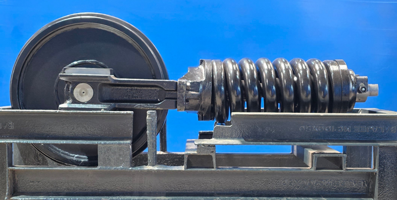A mechanical device with a large black pulley, a coil spring, and metal components mounted on a metallic base against a blue background.