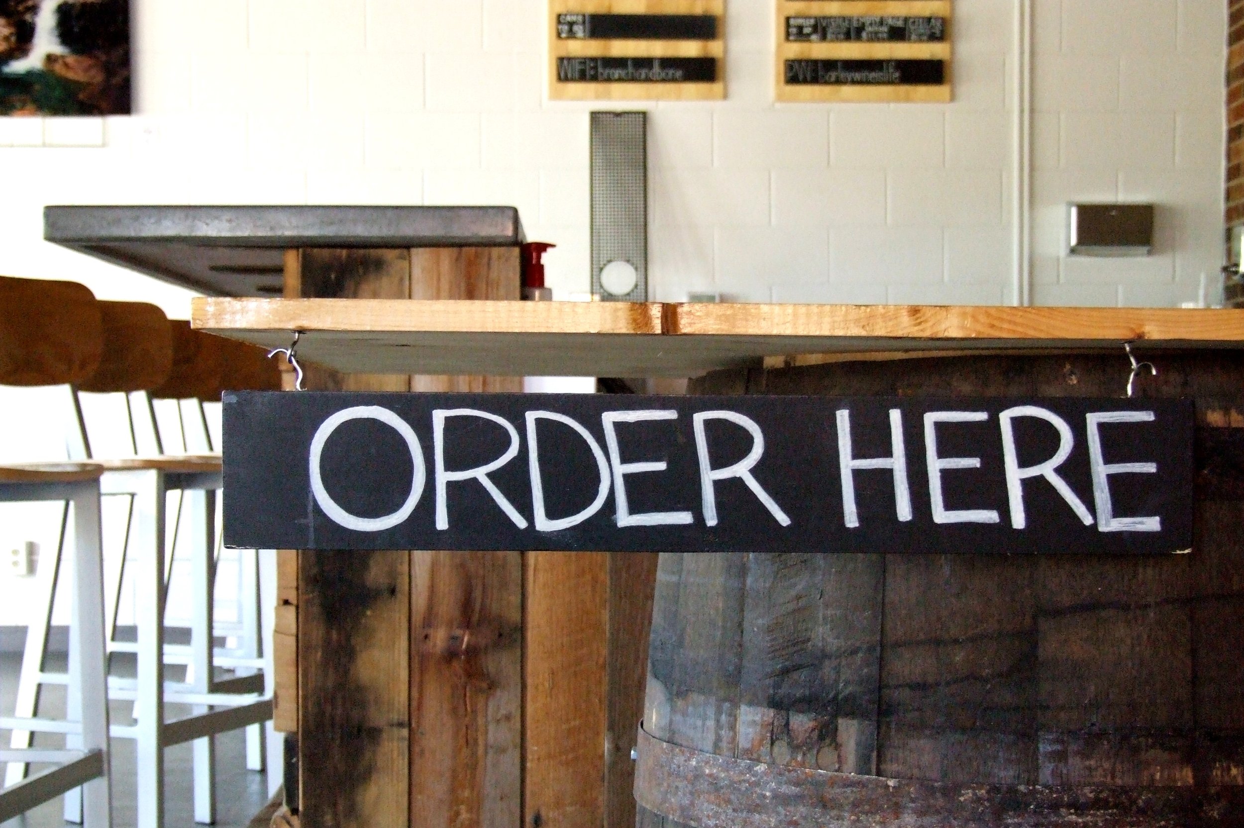 Photo of Order Here sign written in chalk on slate hanging from the end of a bar counter.