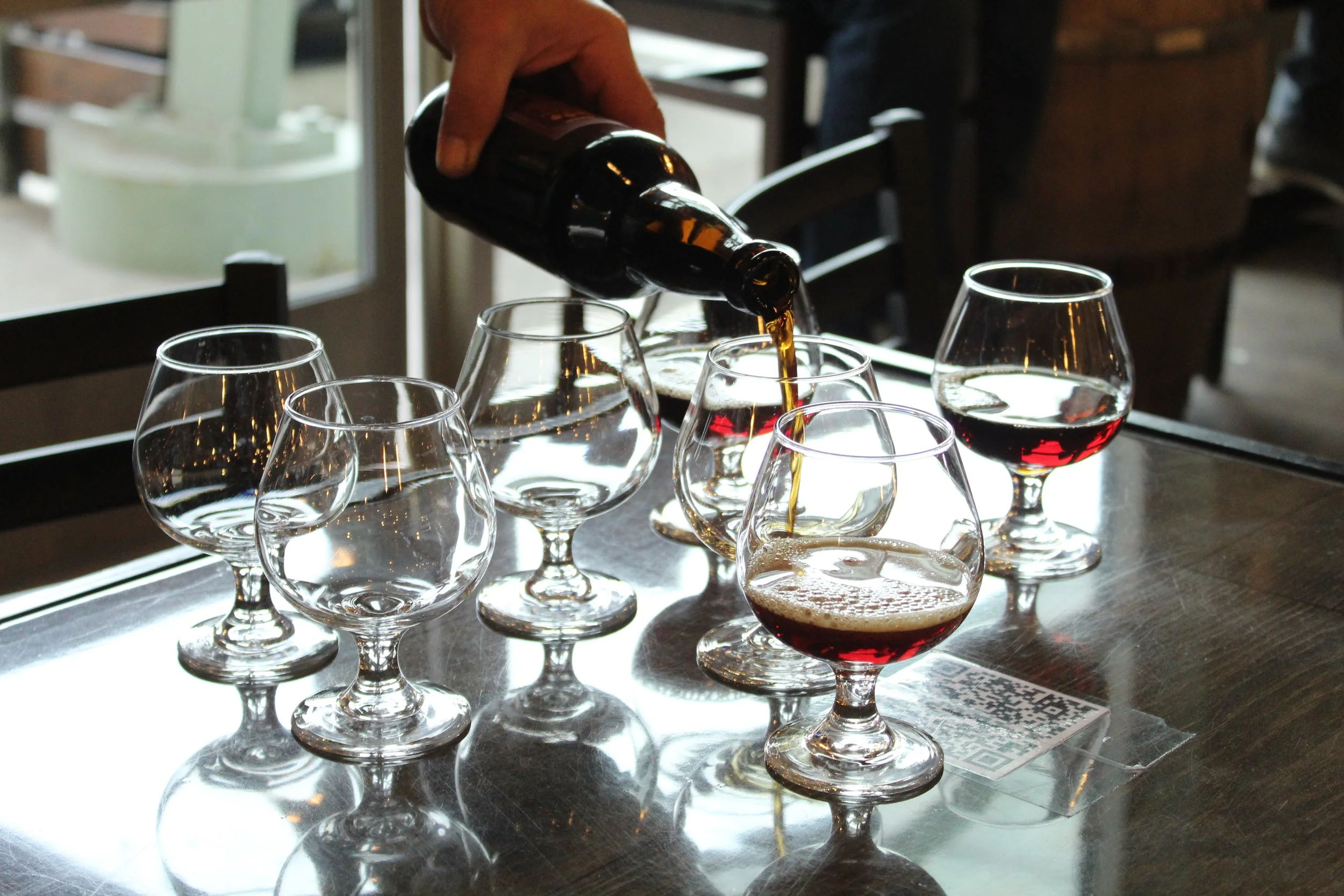Landscape image of multiple snifter beer glasses on a table, some containing an amber-colored beer, with a hand pouring beer into one of the glasses.
