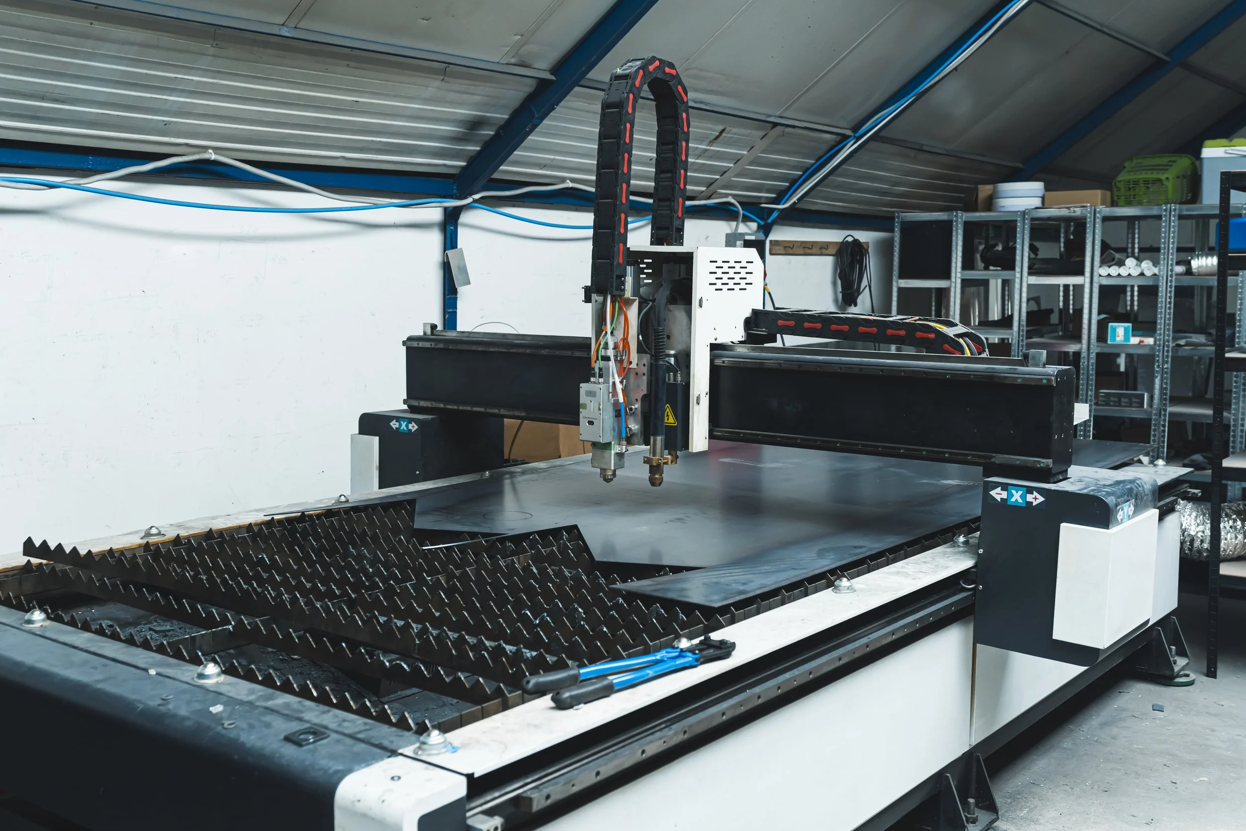 Impresora láser industrial en un taller, con cableado y estanterías con herramientas y suministros en el fondo.