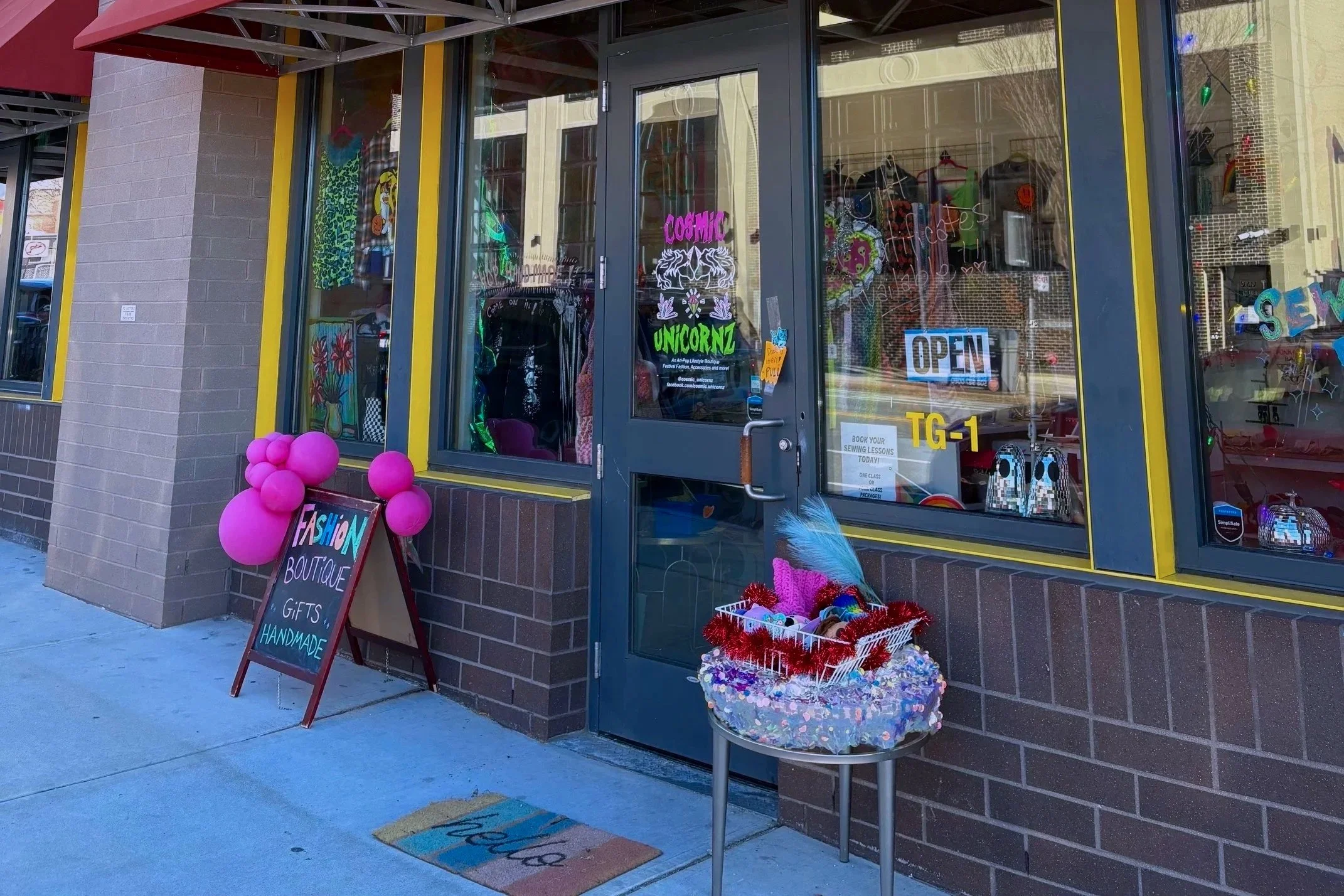 Exterior of a boutique store decorated with pink balloons and a signboard reading 'FASHION BOUTIQUE GIFTS HANDMADE'. The store window displays colorful merchandise and signs, including an 'OPEN' sign. A small table outside holds colorful craft supplies, and a sign on the door advertises sewing lessons.