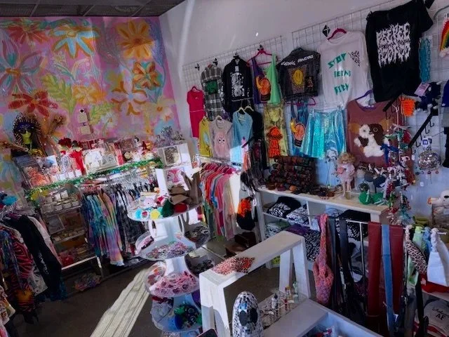 A colorful boutique with a tropical floral mural wall, displaying various clothing items, accessories, and jewelry on racks and shelves.