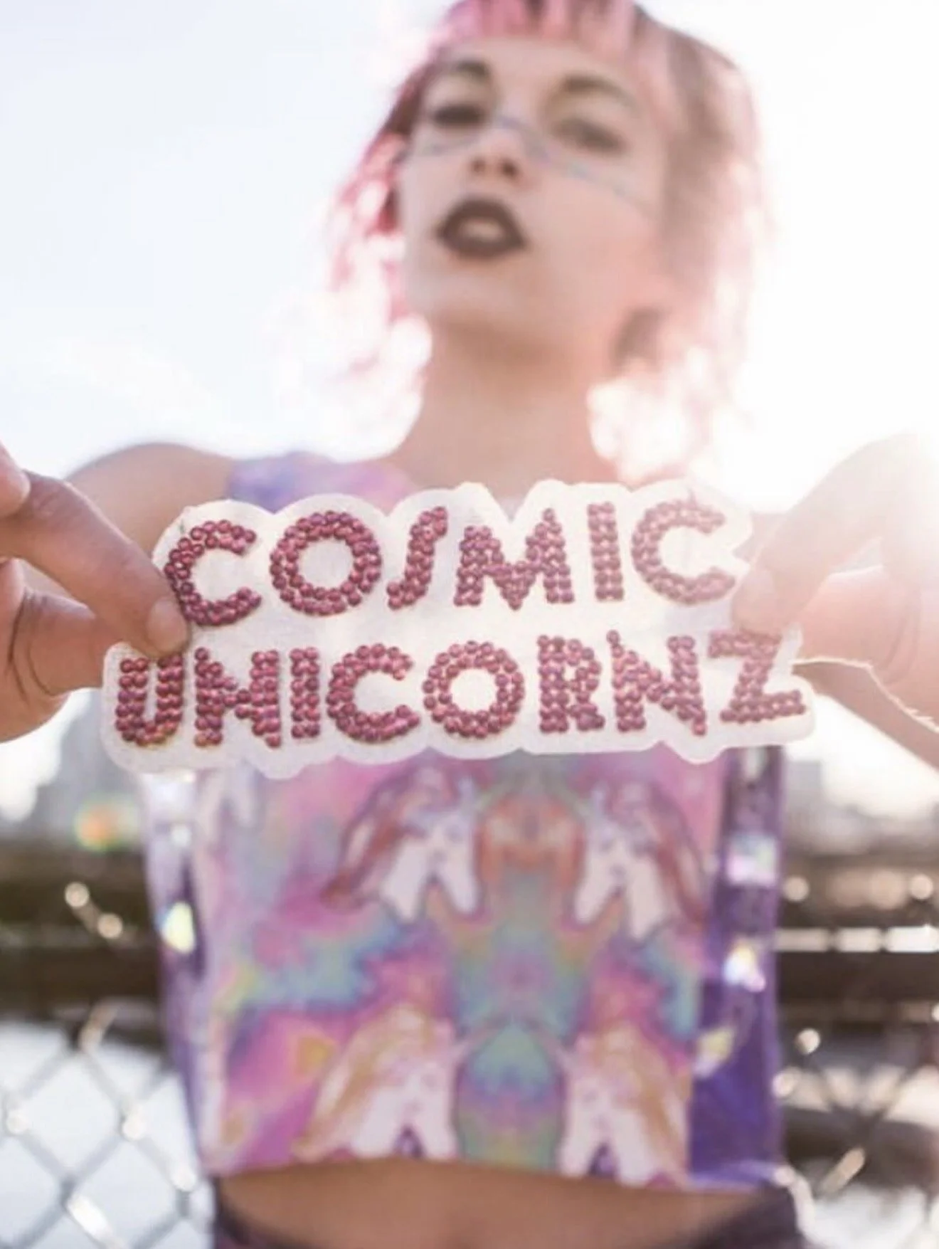 A woman with pink hair and glasses holding a sign that reads 'COJIMIC UNICORN' in pink rhinestones, with sunlight in the background.