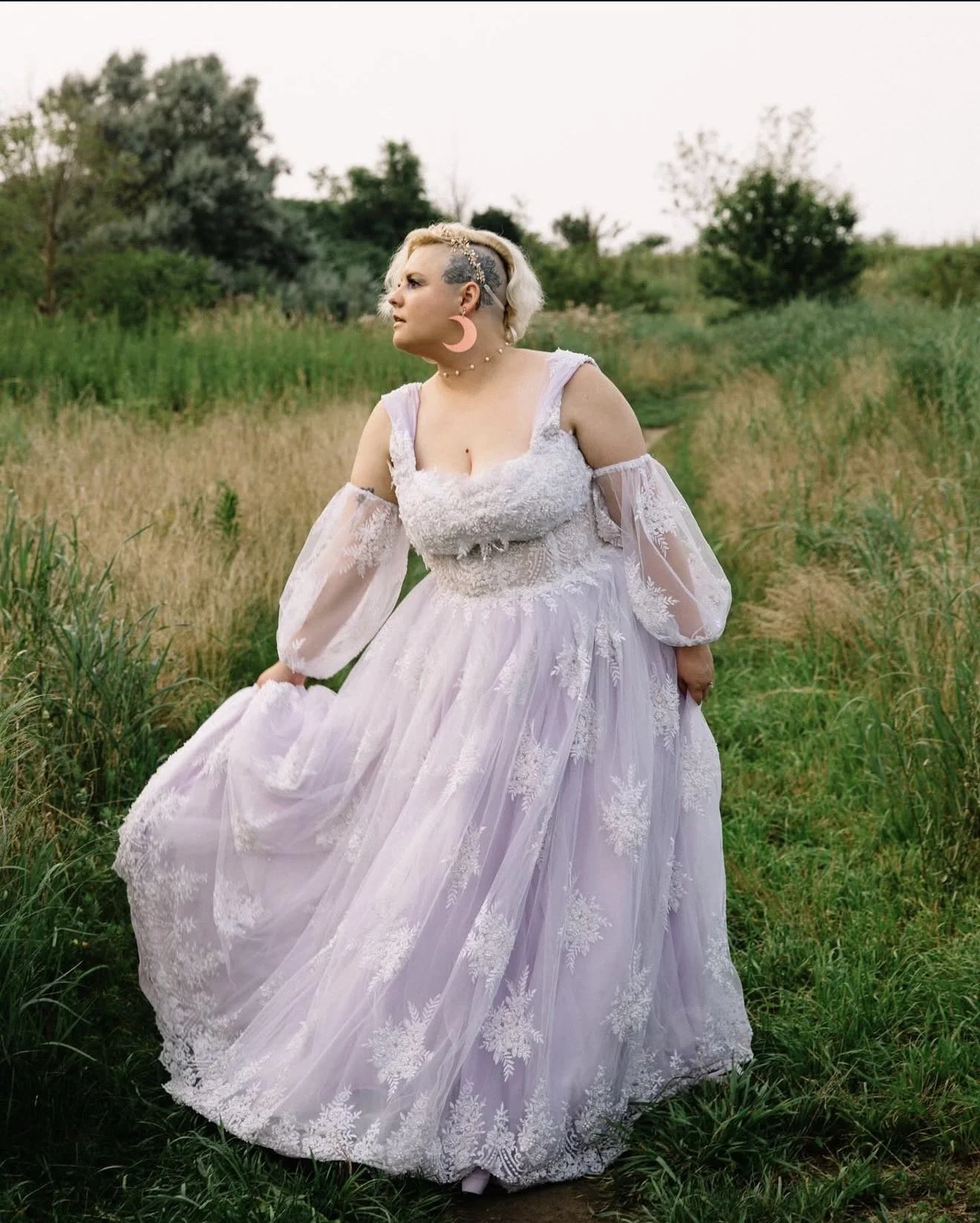 A person in a white lace dress stands in a grassy field with trees in the background, wearing large pink crescent moon earrings and a choker necklace, with short blonde hair styled to one side.