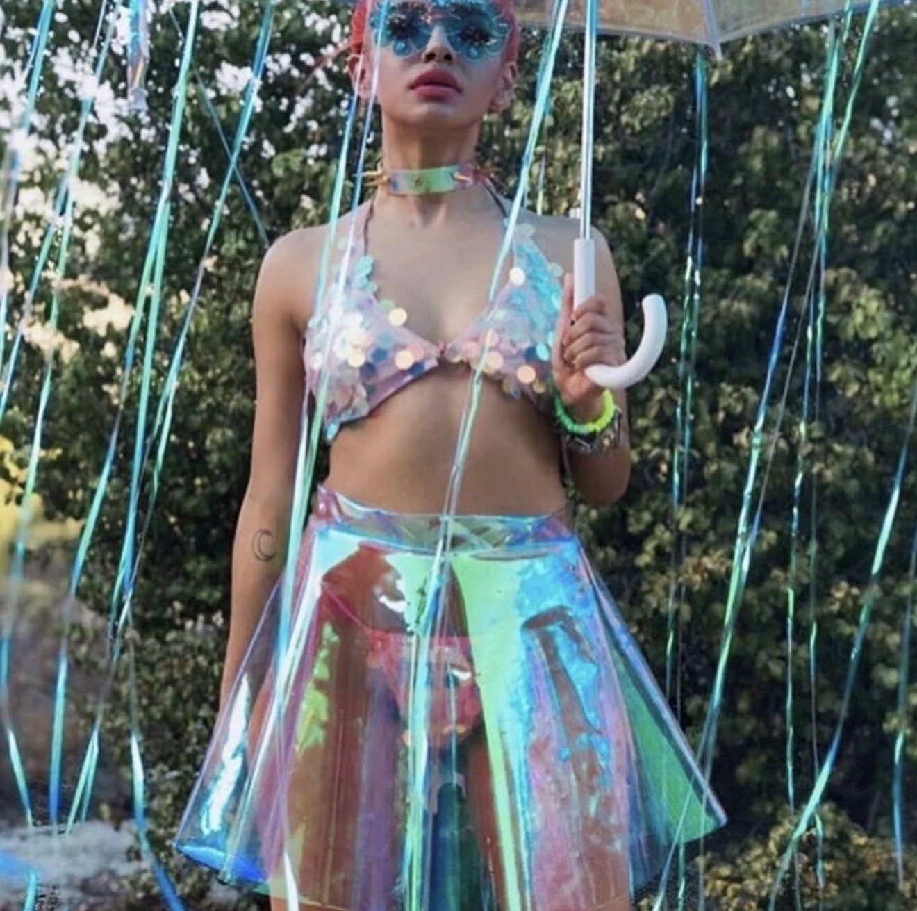 A young woman with pink hair and blue sunglasses holding a transparent umbrella standing outdoors among trees, wearing colorful, reflective clothing including a pastel tank top and iridescent skirt.