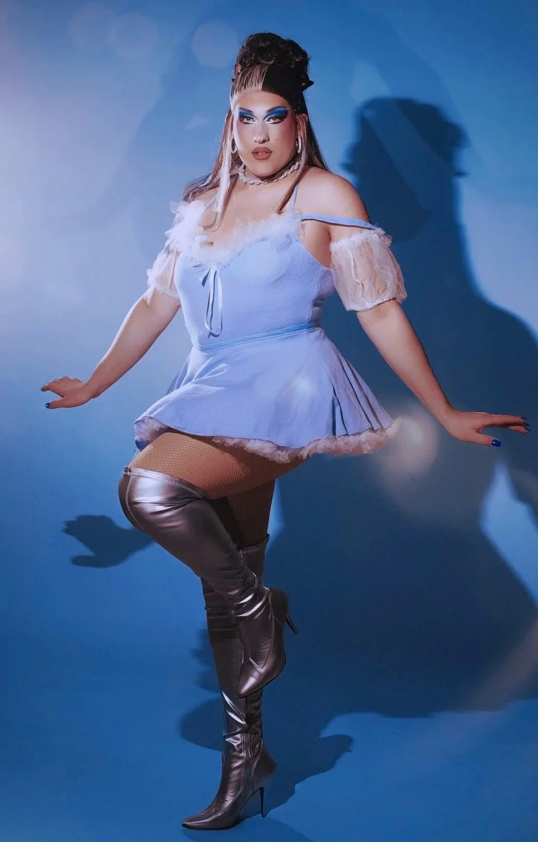 Person dressed in a pastel blue outfit with knee-high silver boots, posing against a blue background with a shadow cast on the wall.