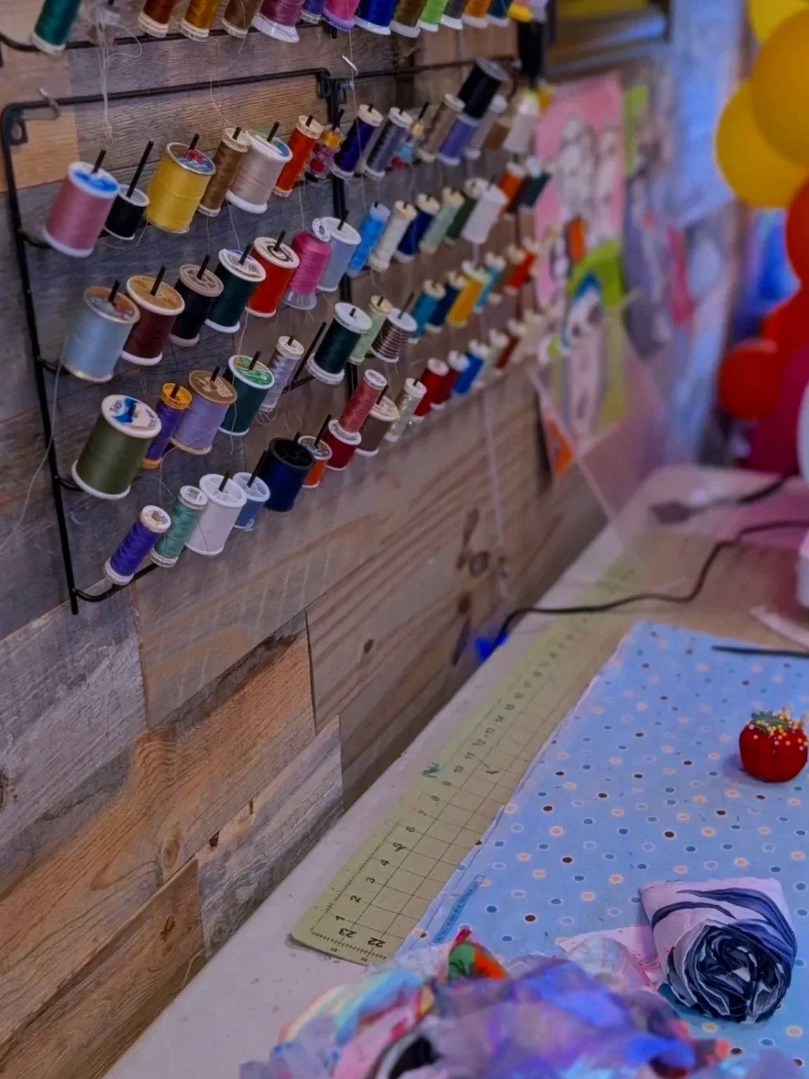 A wall display of colorful sewing threads on spools, with a worktable underneath holding fabric pieces, a cutting mat, and a red pincushion.