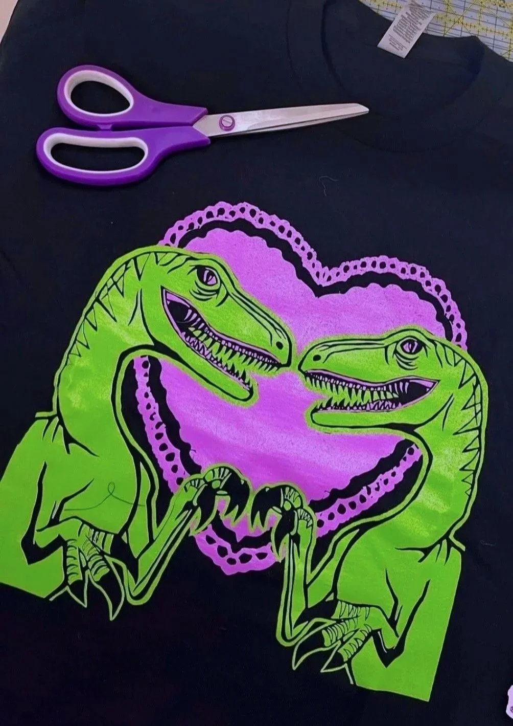 A black fabric with a bright green and purple dinosaur design, a pair of purple scissors, and fabric with a cutting pattern.