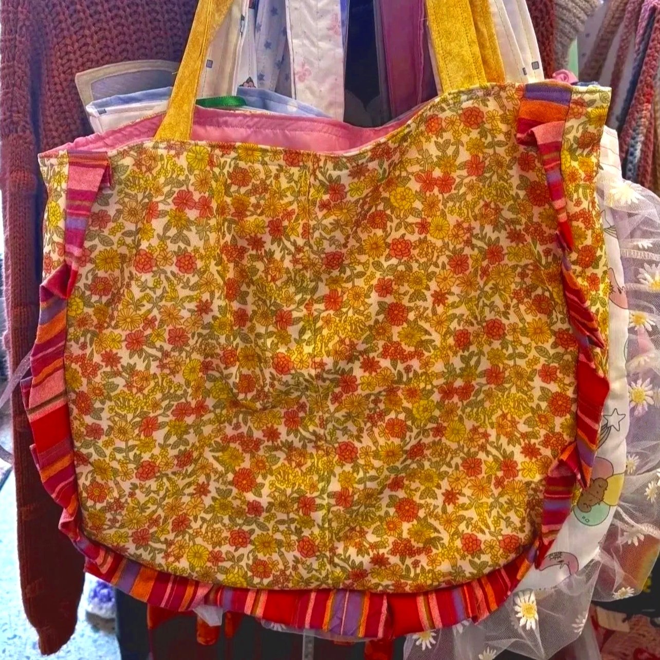 Fabric gift bag with yellow, orange, and green floral pattern, green handles, pink lining, and ruffled edges, displayed among other colorful textiles.