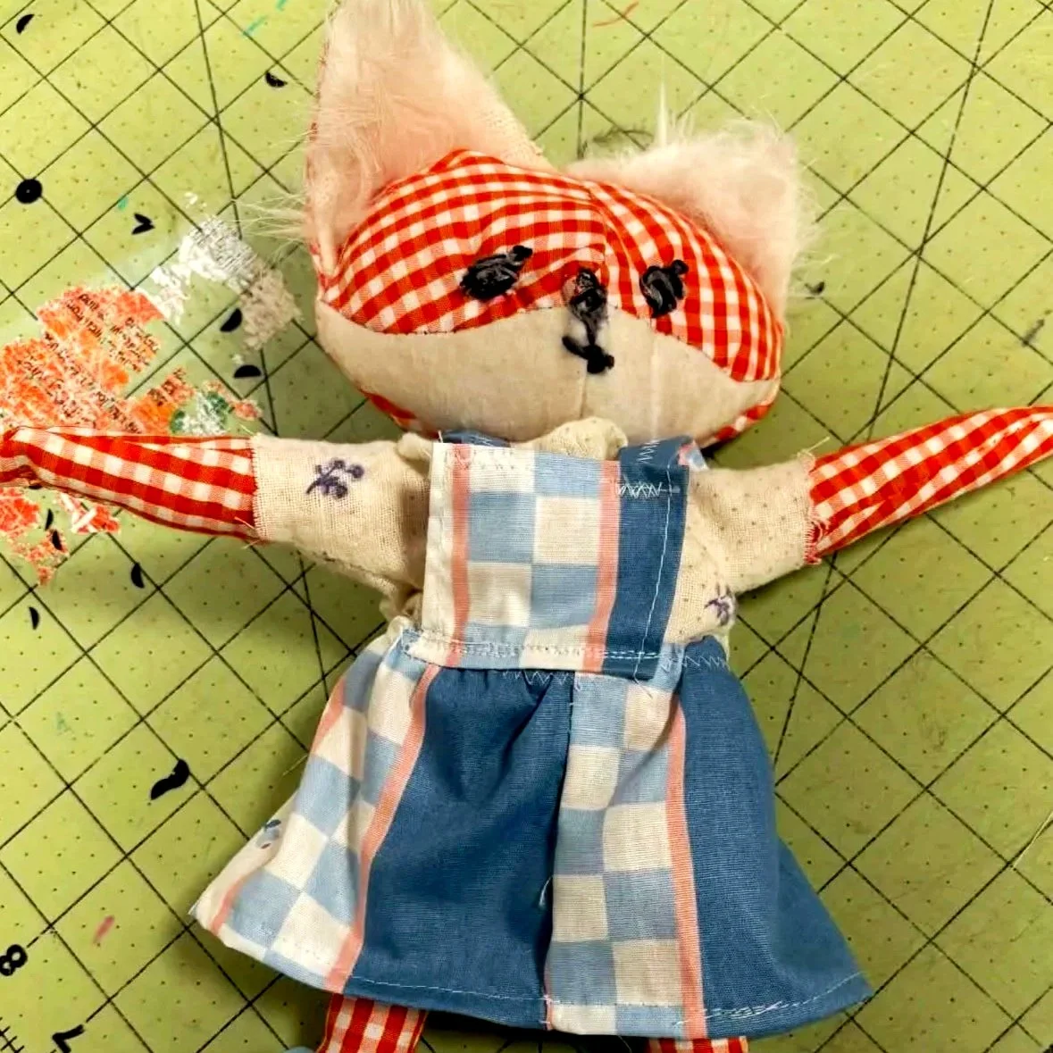 A handmade cloth doll with a fabric face and limbs, dressed in a patchwork dress, embroidered with lavender flowers, lying on a green cutting mat with black measurement lines and black markings.