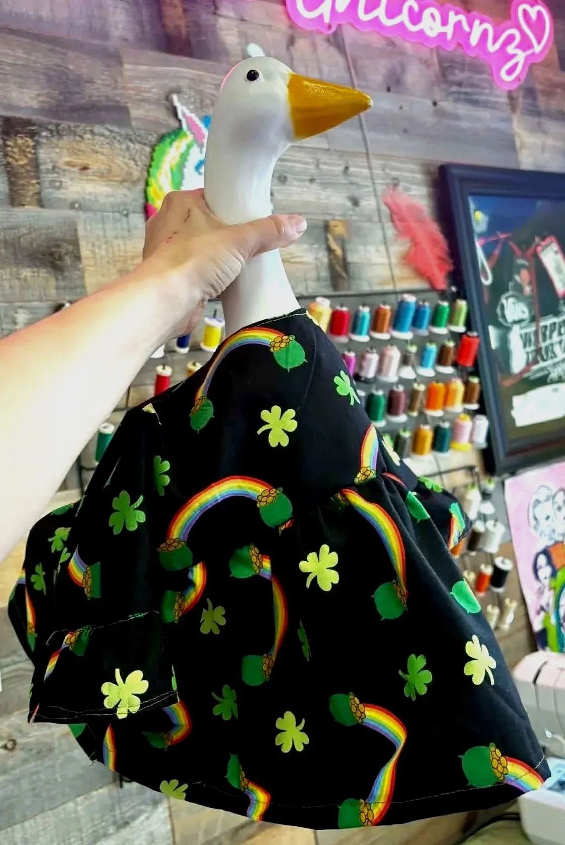 A stuffed goose toy wearing a black dress with green shamrocks and rainbow patterns, held by a person's left hand in front of colorful threads and a framed sign in a craft store.