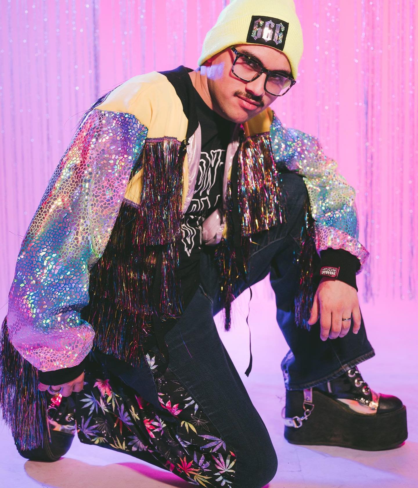 A person dressed in colorful, eclectic fashion posing in front of pink shiny backdrop with hanging tinsel.