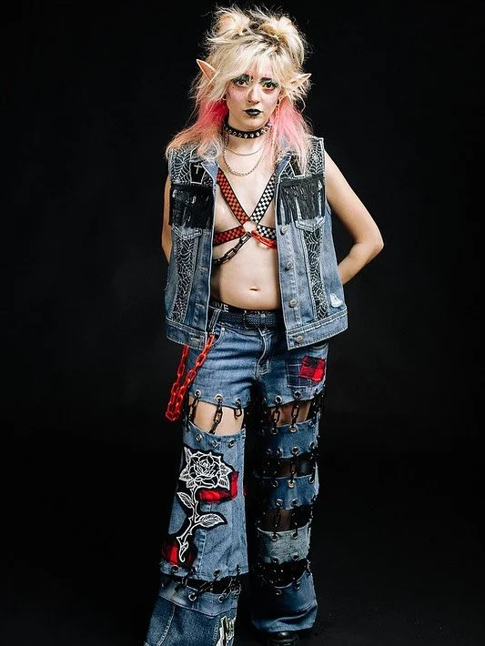 A young woman with blonde spiky hair and pink streaks, wearing dark makeup and Goth style clothing, standing against a black background.