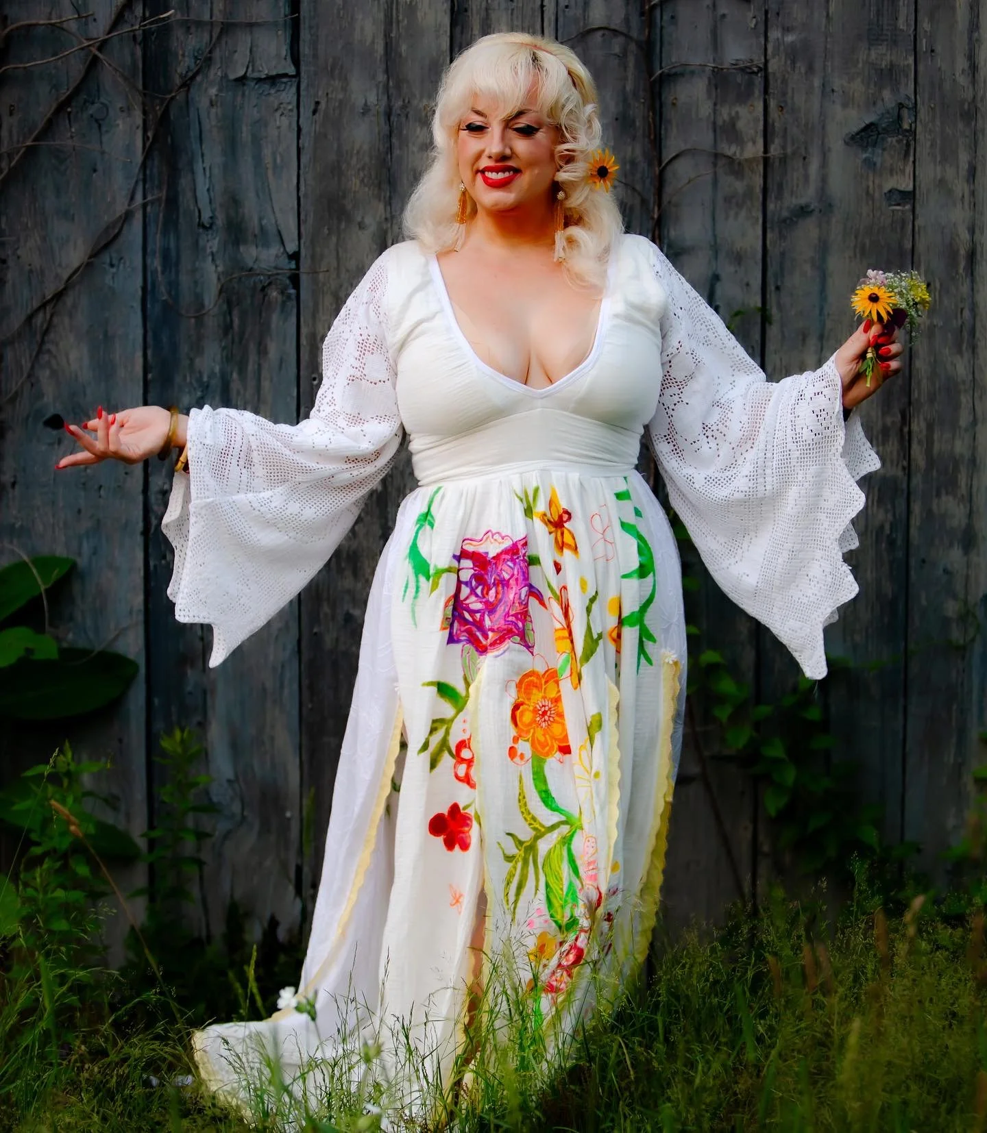 Woman with blonde hair wearing a white dress with colorful floral embroidery, standing outdoors in front of a wooden fence, holding flowers in both hands, smiling.