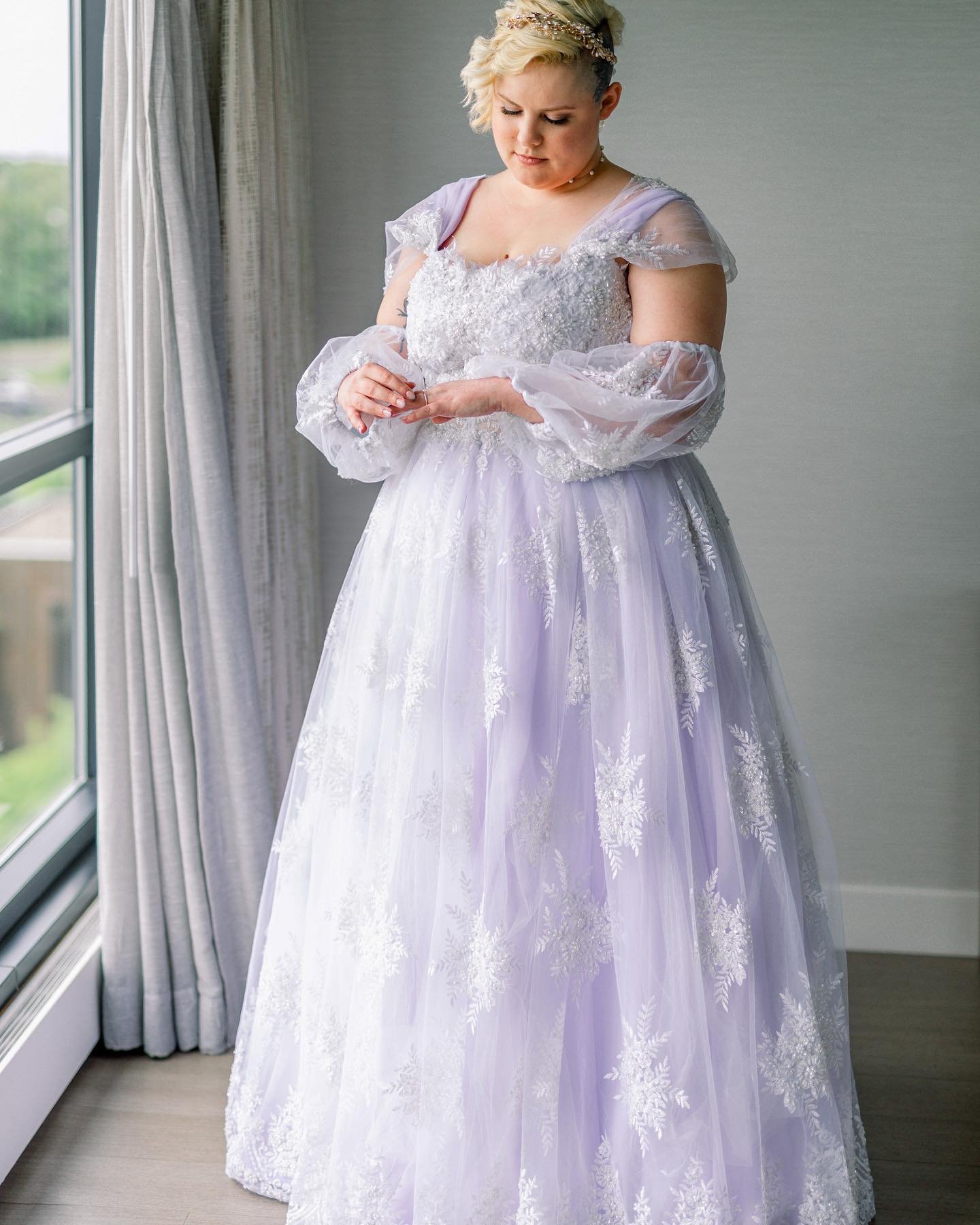 A woman in a white, ornate wedding dress with sheer sleeves and lace details, standing by a window inside a room.