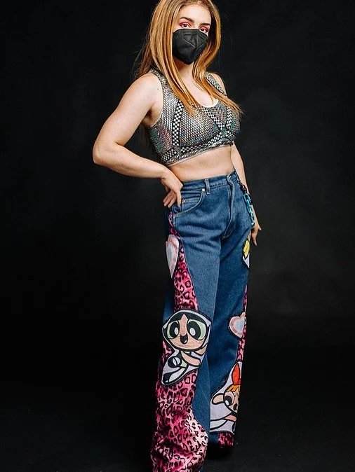 A woman with long red hair wearing a black face mask, a patterned crop top, and colorful jeans with cartoon characters design, standing against a black background.