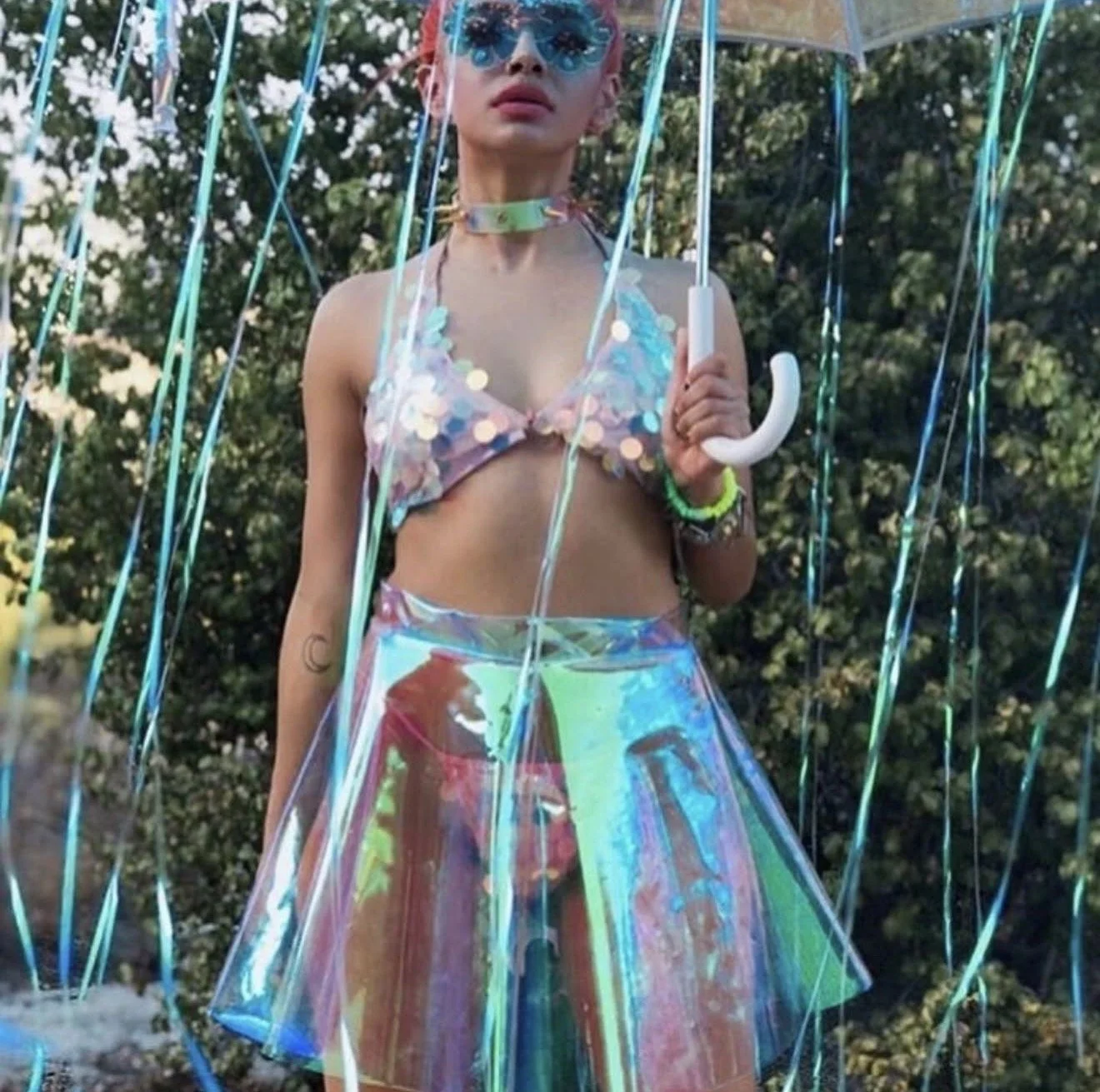 A young woman wearing a colorful, reflective skirt and a iridescent crop top with sequins, holding a clear umbrella with rainbow reflections, standing outdoors surrounded by blue and teal streamers.