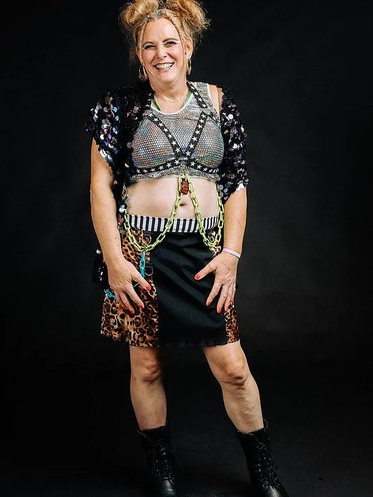 Woman smiling, wearing a colorful, eclectic outfit with a sequined top, leopard print and striped skirt, accessories, and black boots.