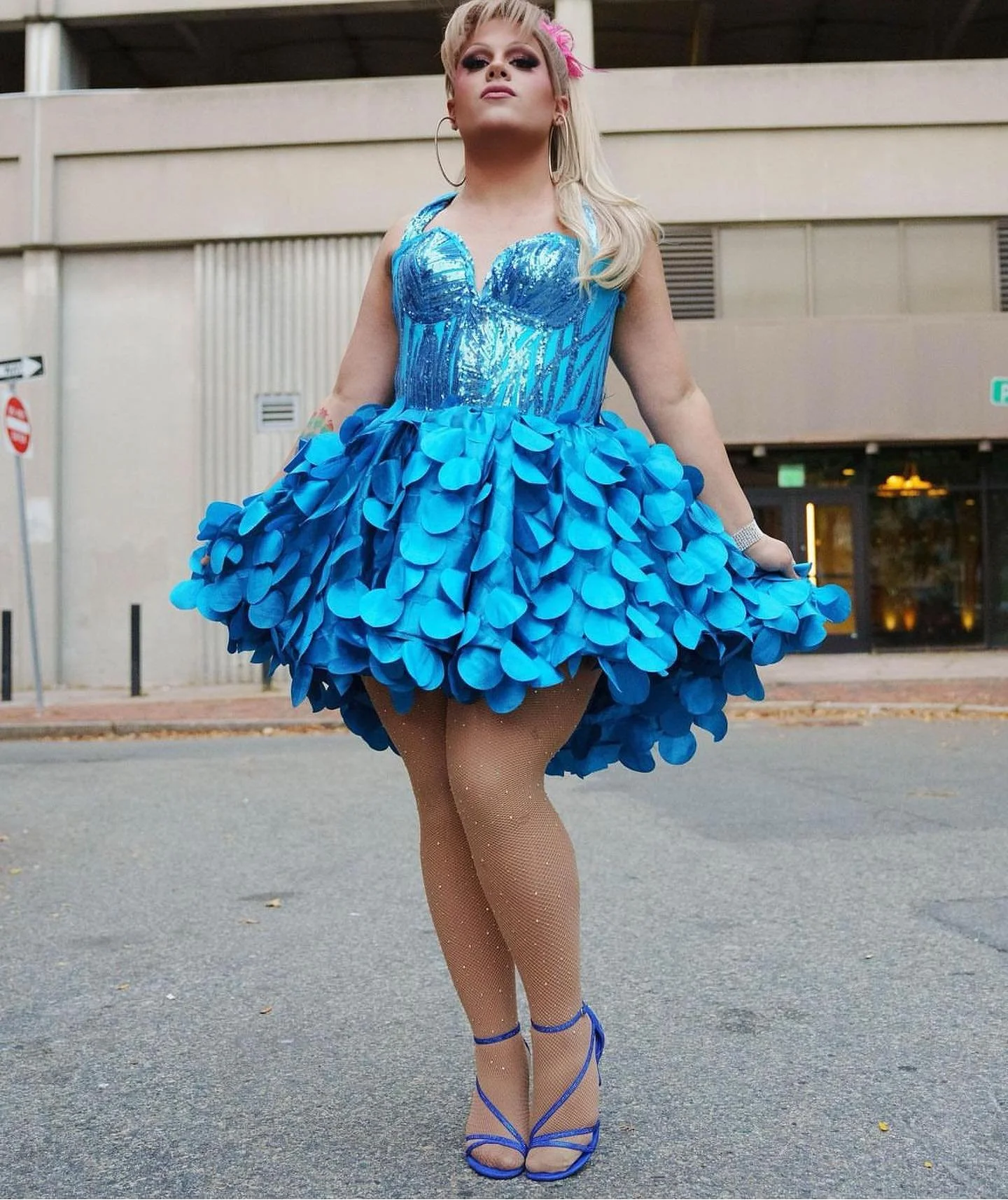 Person dressed in a sparkling blue dress with a fluffy skirt, fishnet stockings, and matching blue high heels, standing outdoors on a city street.