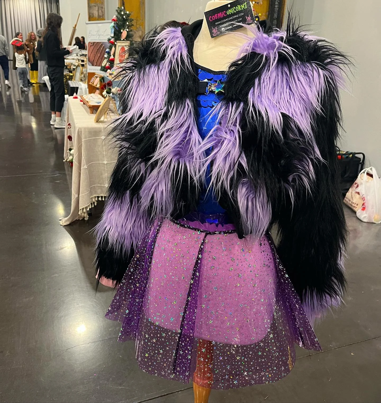 Mannequin dressed in a colorful, sparkly fairy costume with a purple glittered skirt, an iridescent blue top, and a large faux fur coat with black and purple feathers, displayed in an indoor setting with people and tables in the background.