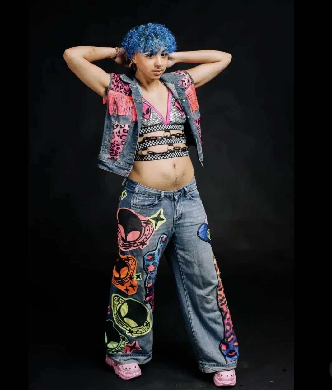 Young woman with bright blue curly hair wearing a colorful patterned jacket, a glittery top, distressed jeans with colorful patches, pink Crocs, and posing with arms behind her head against black background.