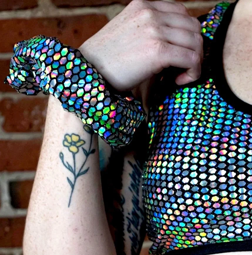 A person with tattoos shows their arm while wearing a holographic, honeycomb-patterned bracelet and top, with a brick wall background.