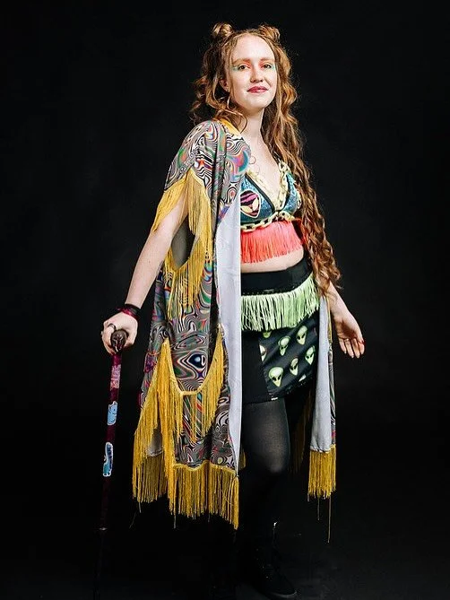Woman with long, curly red hair wearing a colorful, fringed, bohemian style outfit, holding a walking cane, standing against a black background.