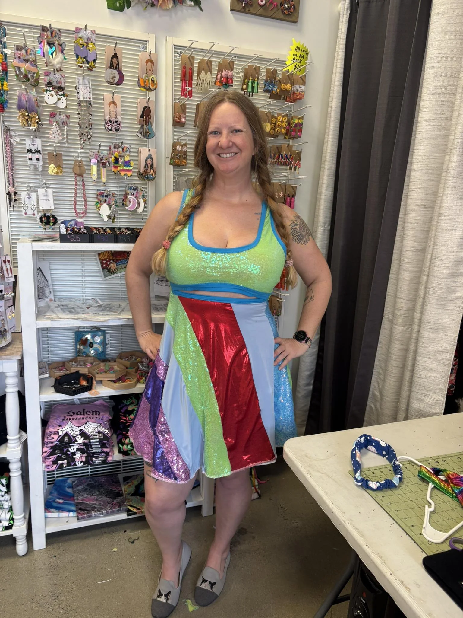 A woman with braided hair smiling stands inside a store wearing a colorful, patchwork dress and grey slippers with black and white cat faces. Behind her are display racks with various jewelry and accessories.
