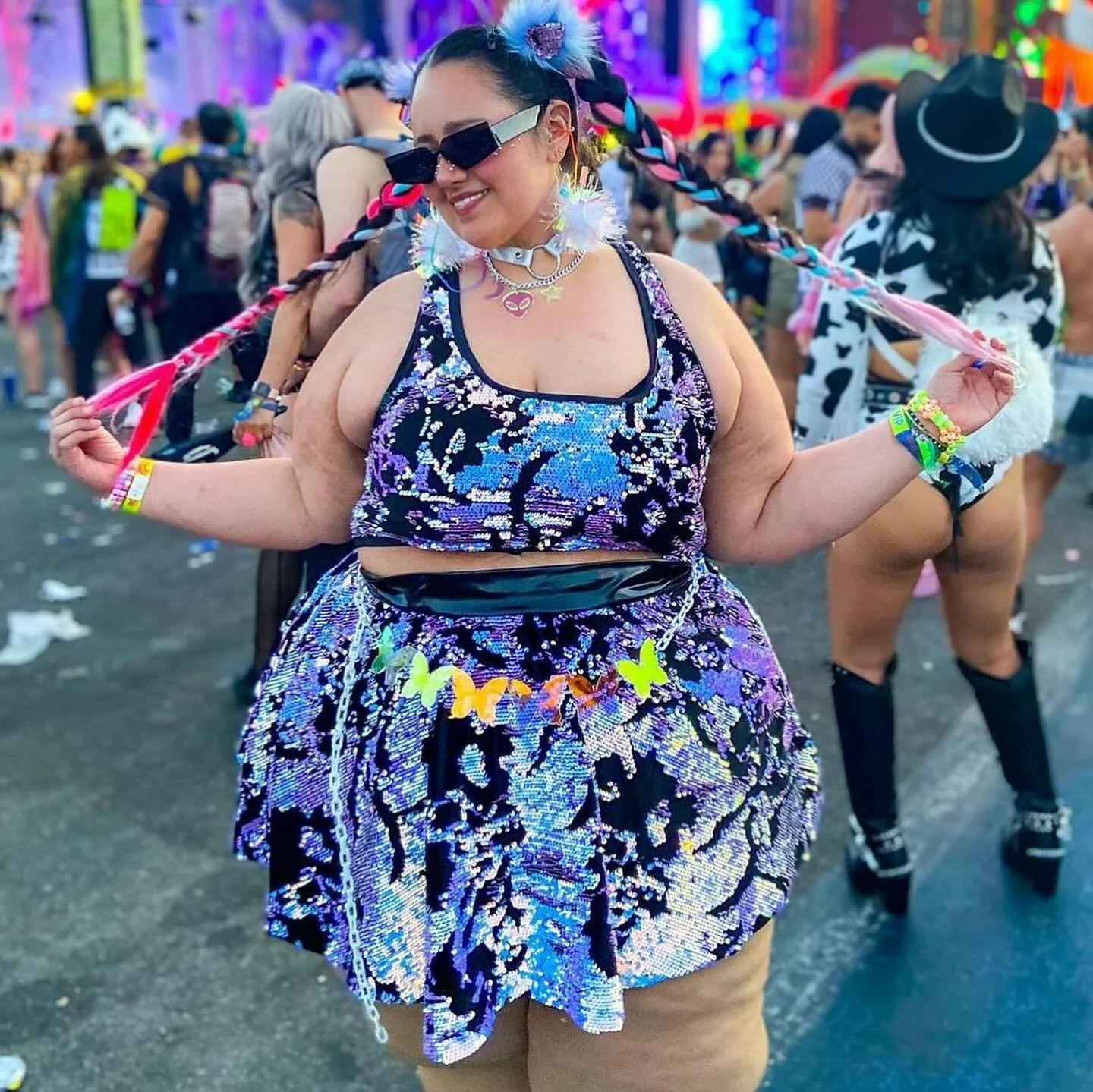 A woman at a festival or pride event wearing a colorful, reflective sequined outfit with butterfly accessories and large sunglasses, smiling while holding her braided hair. In the crowd background, other dressed-up festival-goers are visible.