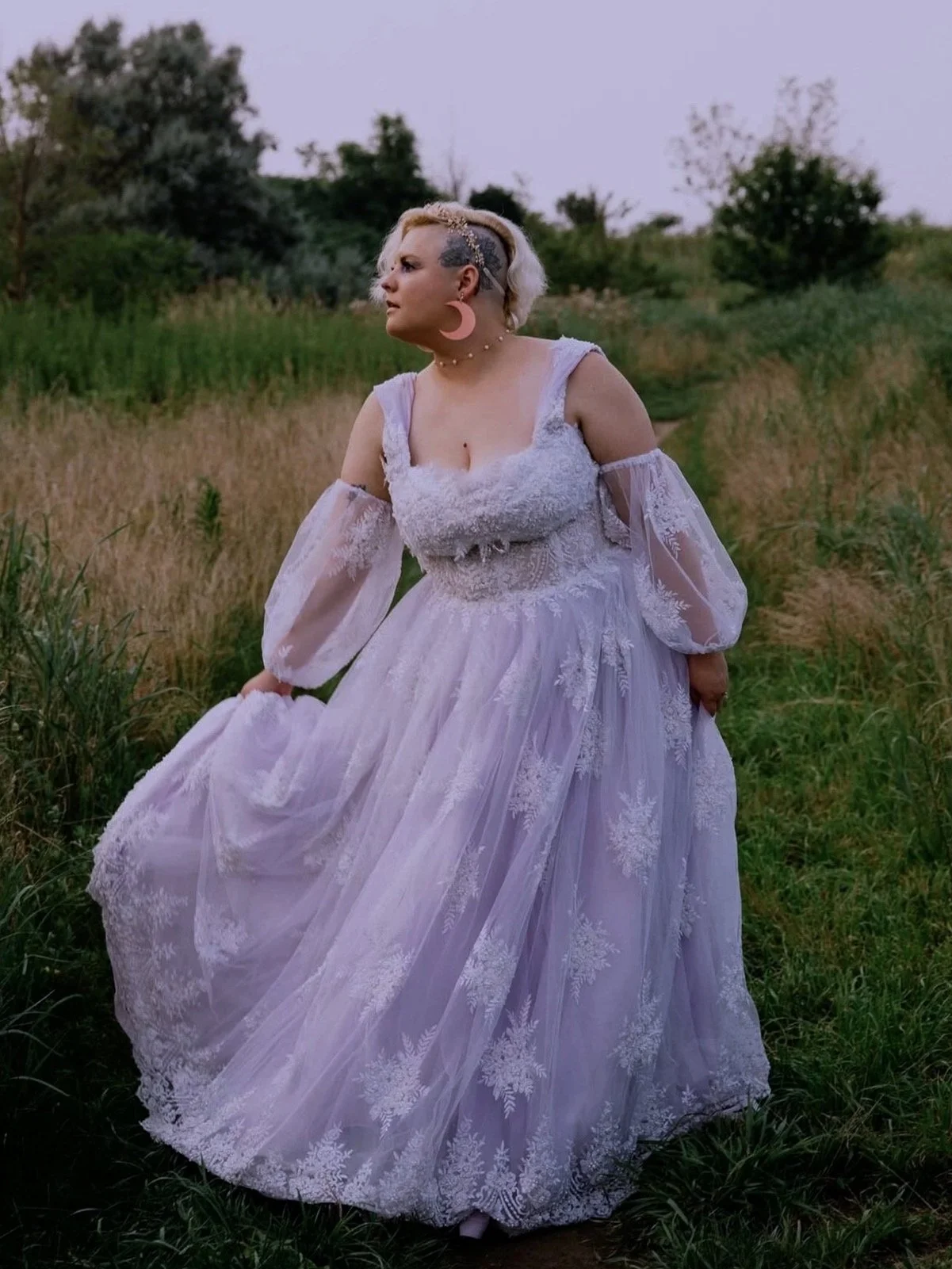 A woman wearing a light purple lace gown with sheer sleeves standing in a grassy field with trees in the background.